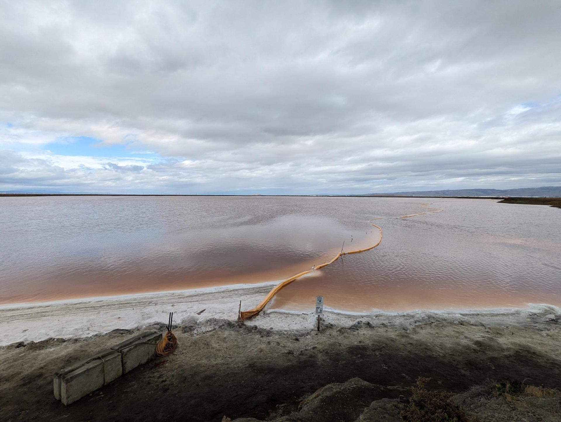 Overcast sky above a wide, reddish-brown body of water with a yellow boom.