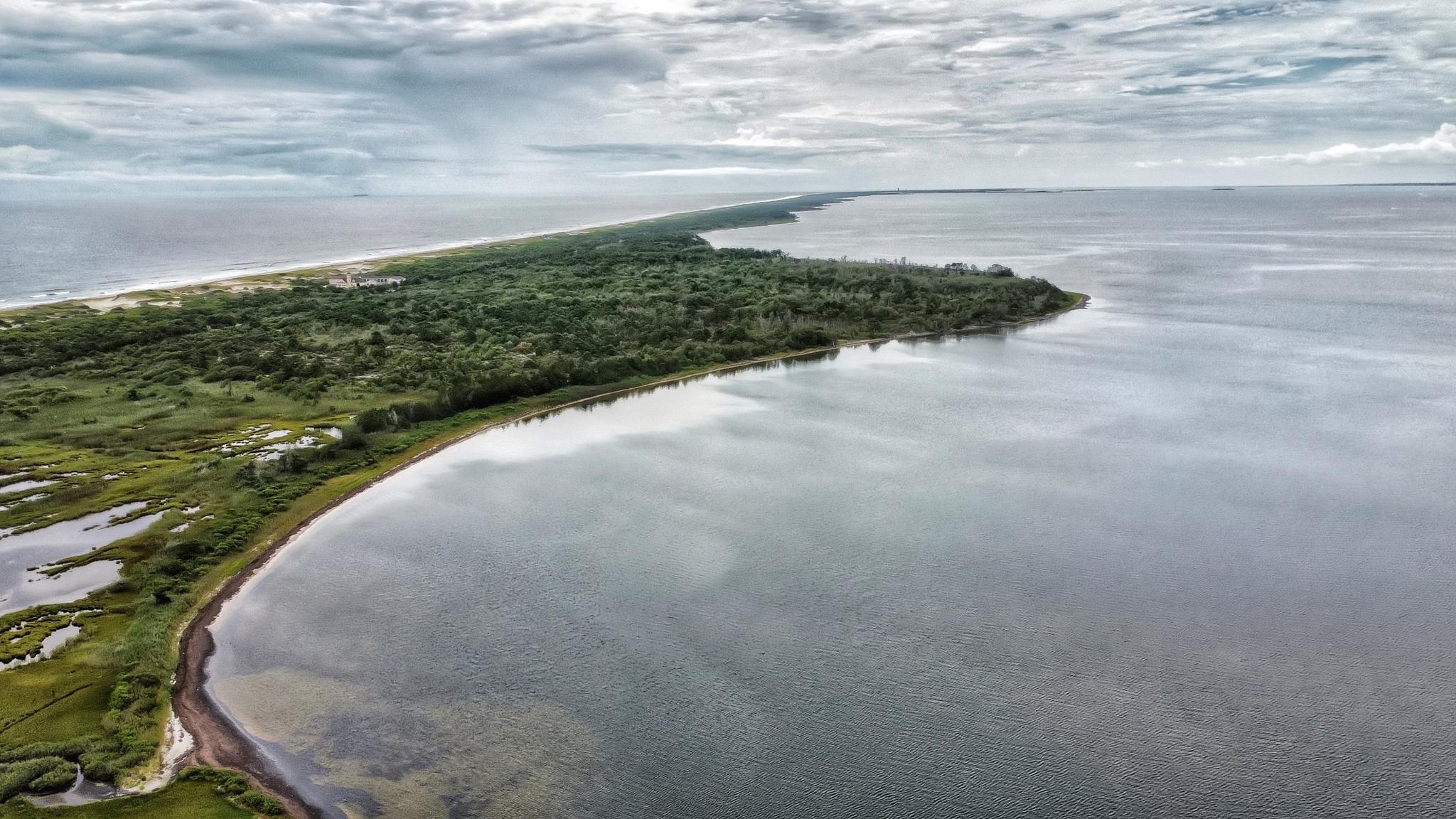 Aerial view of a long, green landmass surrounded by calm water under cloudy skies.