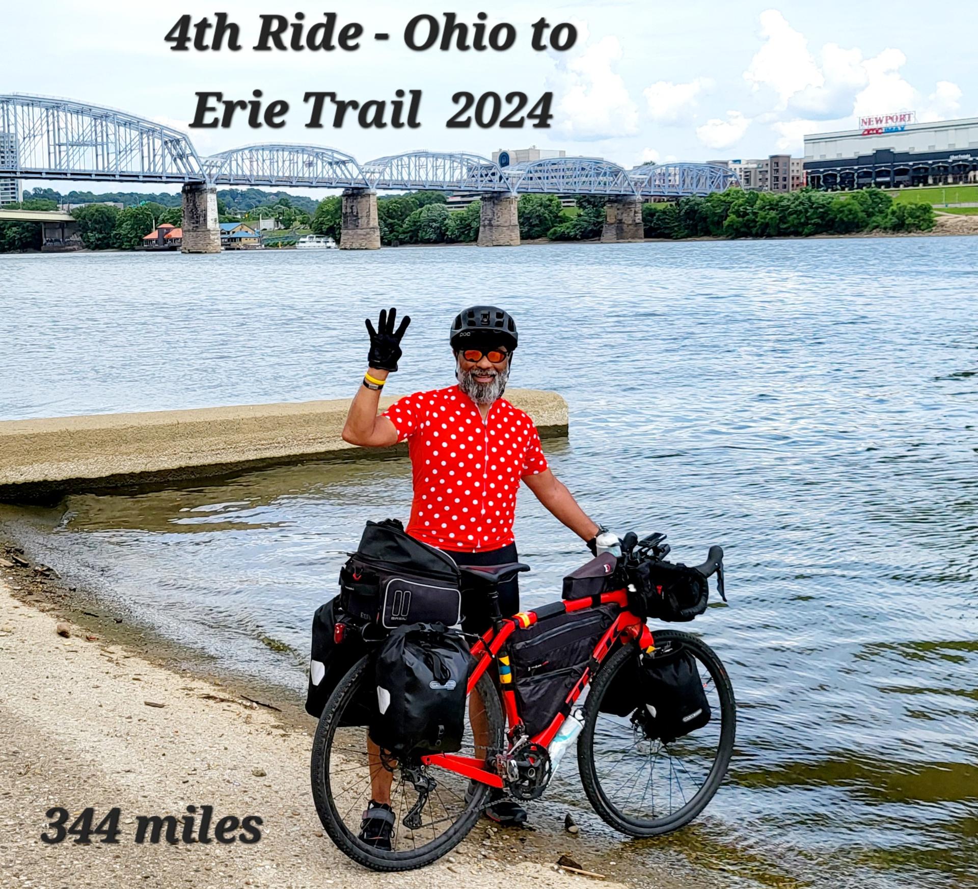 A cyclist with a loaded bike stands by a river, waving, under a bridge.