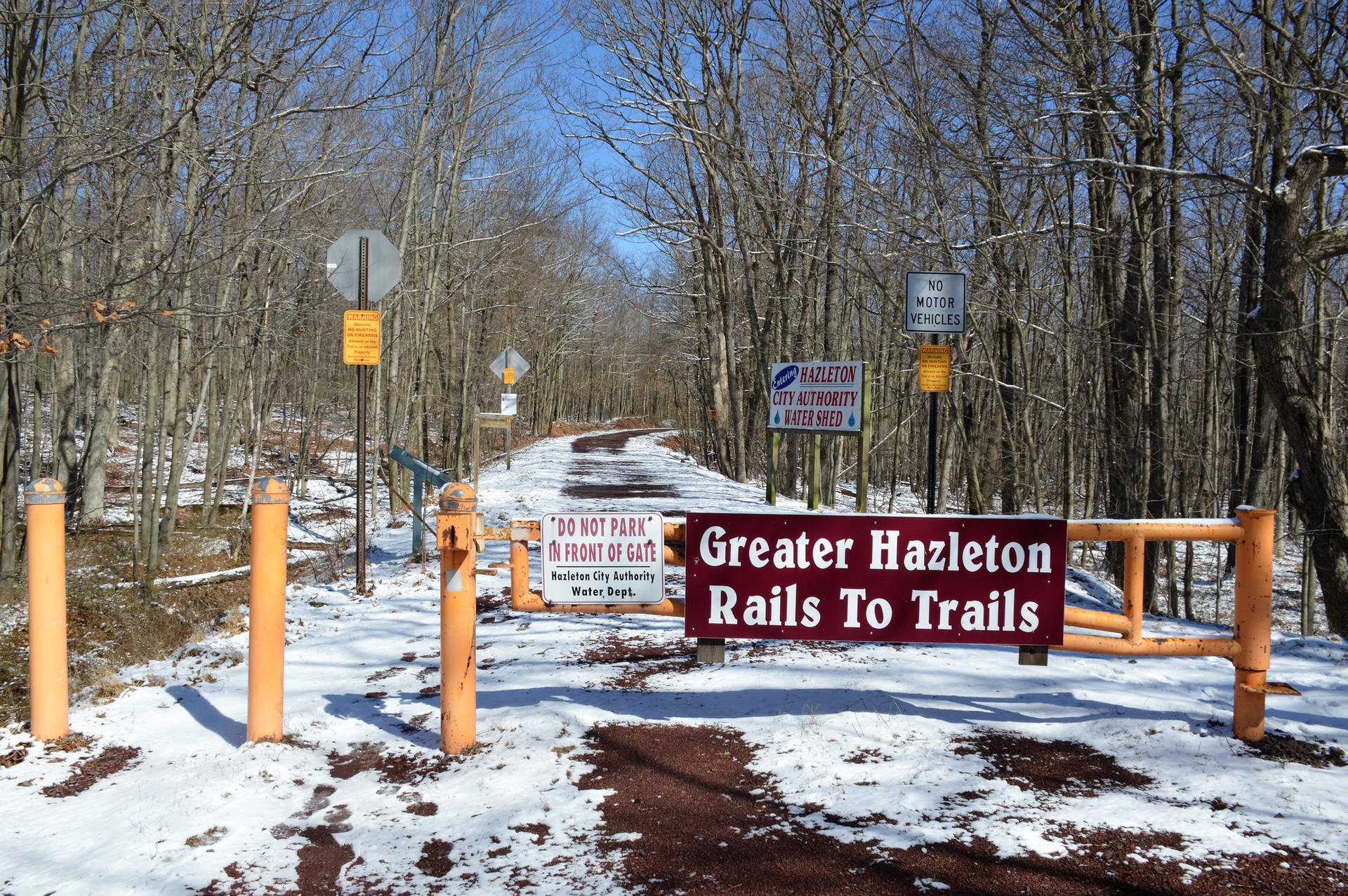 Snowy trail entrance with a gate and signs in a bare winter forest.