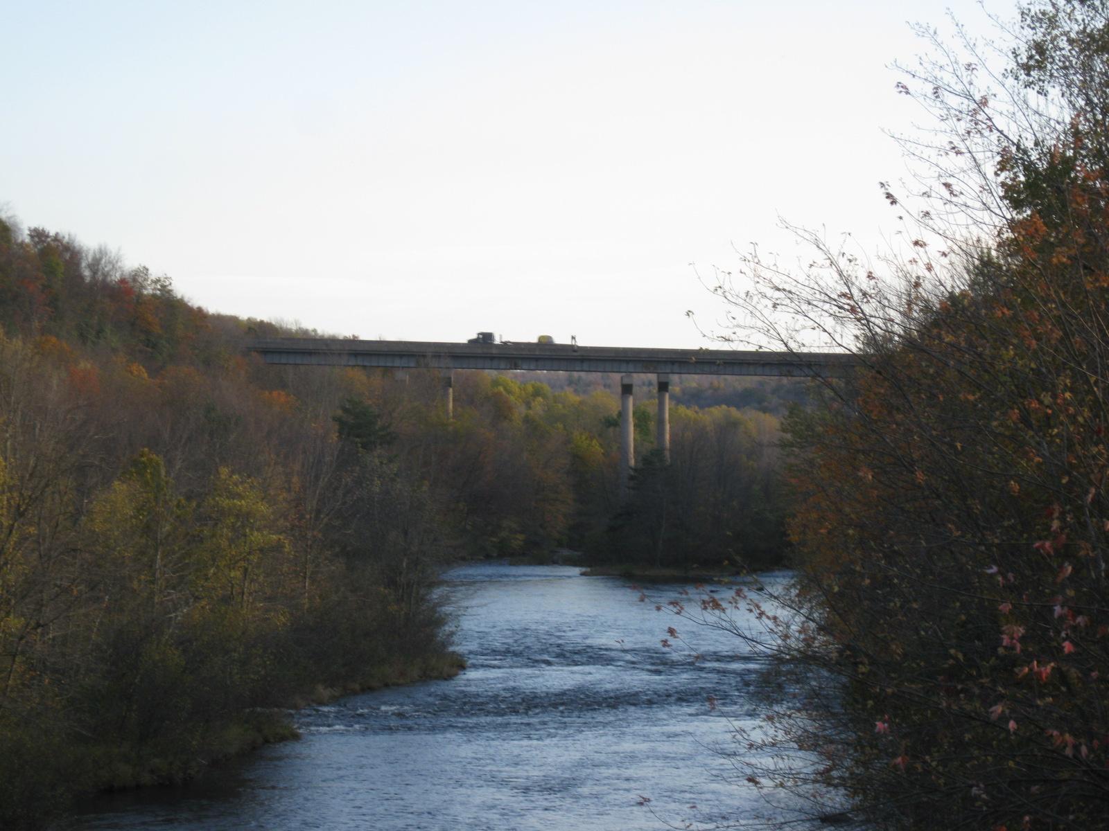A river flows through autumn trees under a distant highway bridge.