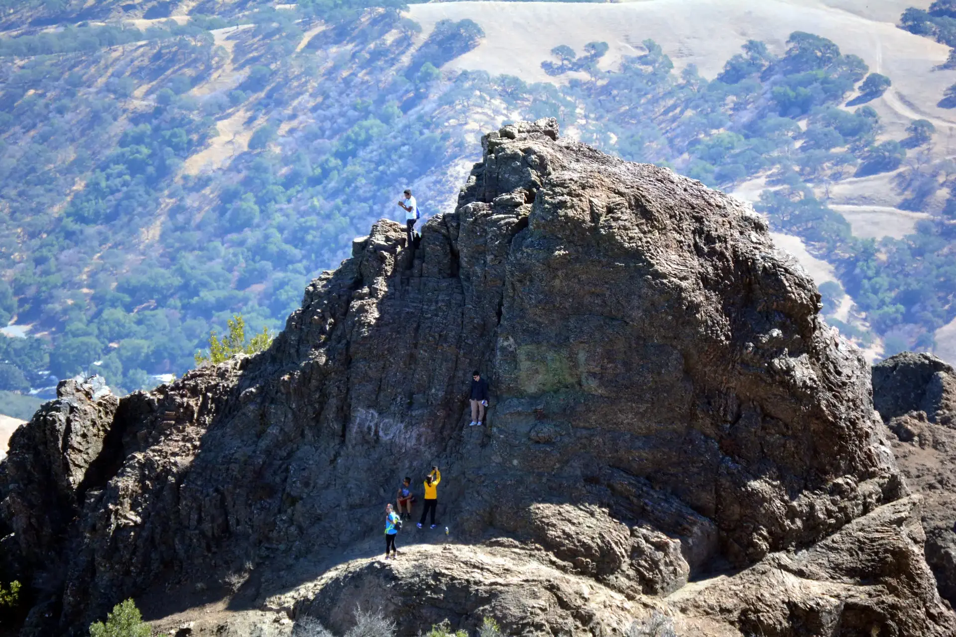 Hikers stand on a jagged rocky peak overlooking green valleys.