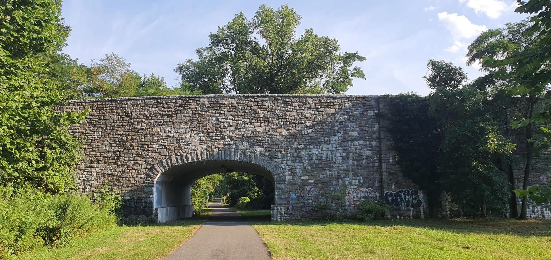 Large stone arch bridge over a paved path, surrounded by green trees.