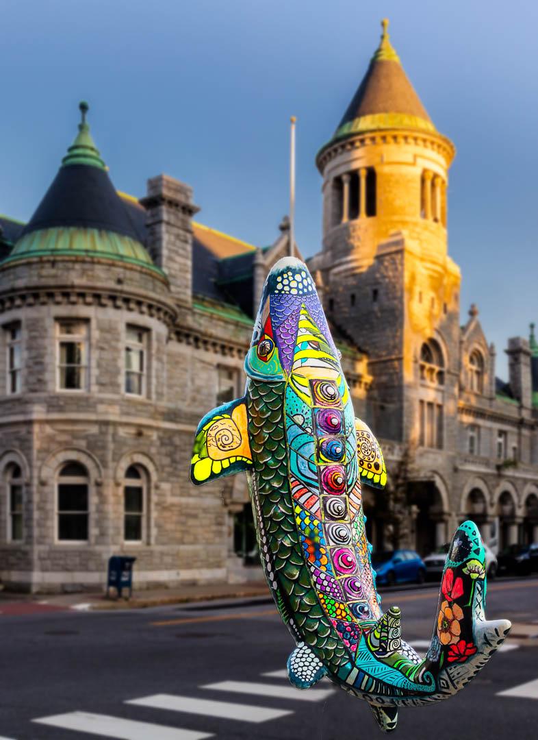 A vibrant, patterned fish sculpture stands on a street with old stone buildings.