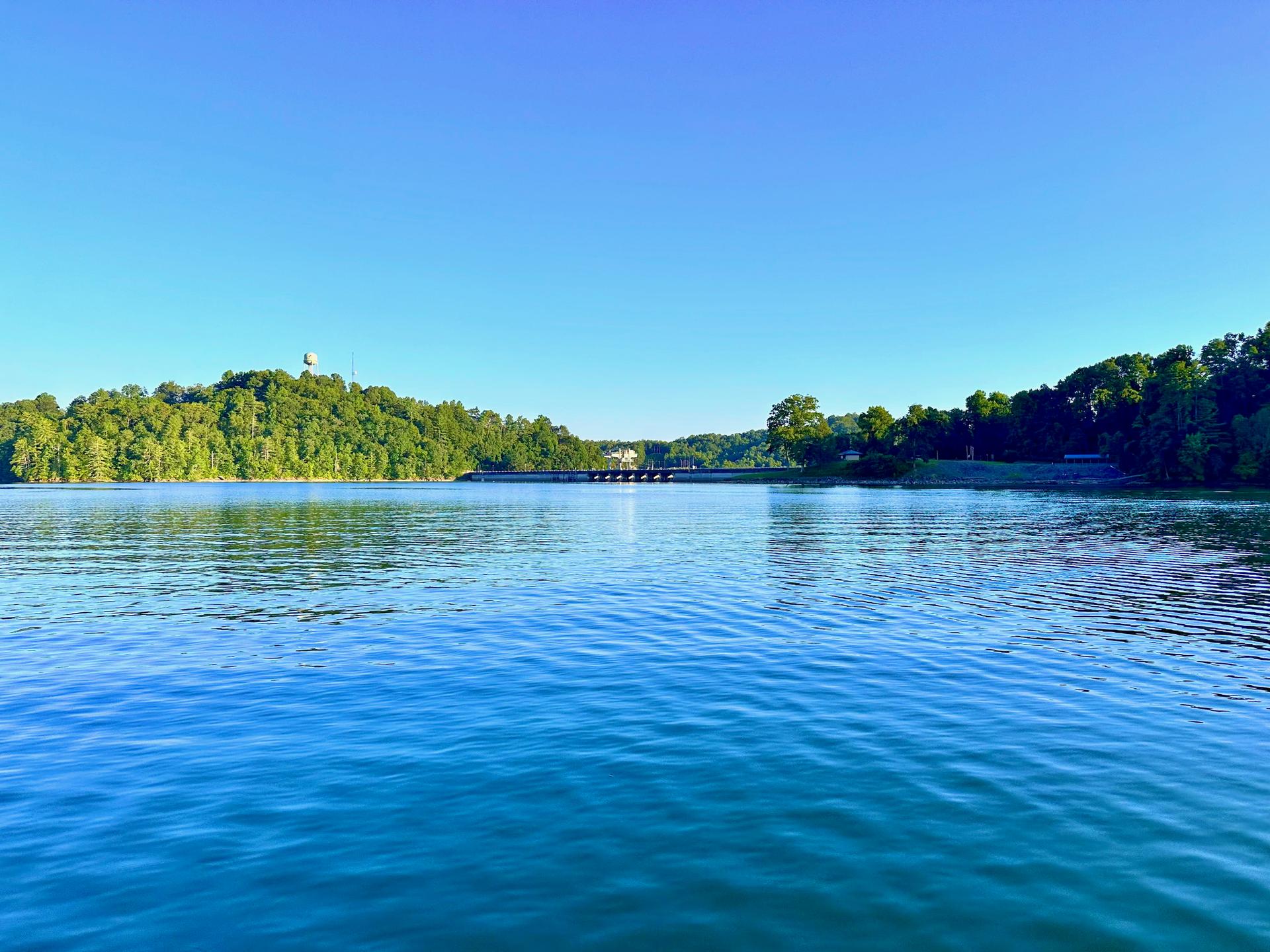 Expansive blue water, green forested hills, and a distant structure under bright sun.