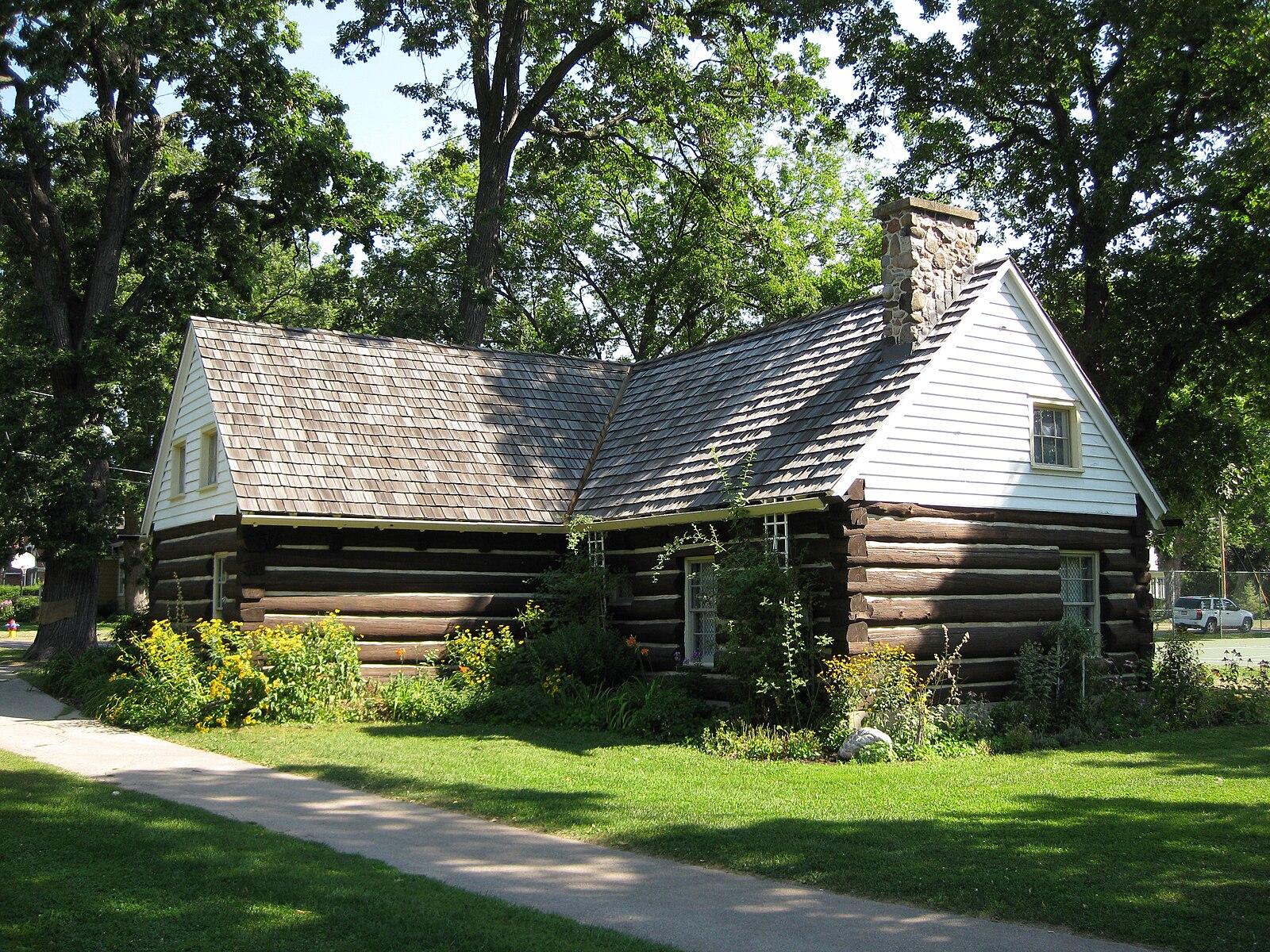 An old log cabin with a shingled roof sits among green trees.