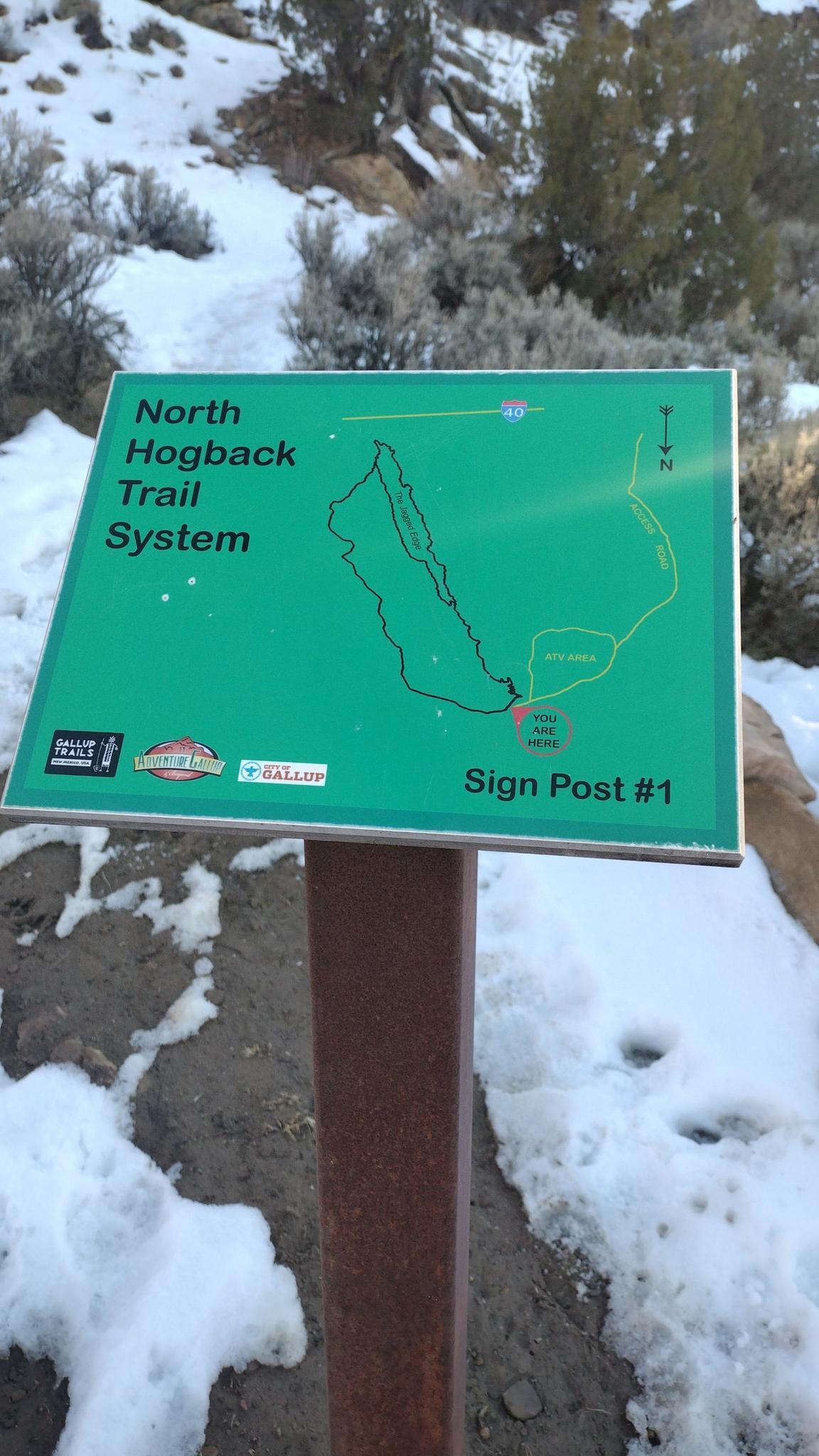 Green trail map sign on a snowy path with sparse vegetation.