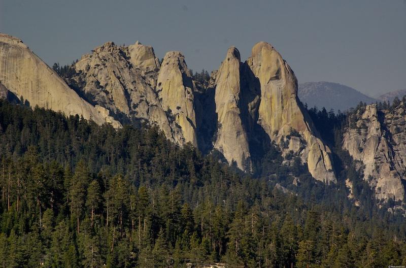Towering rock formations rise above a dense green forest under a clear sky.