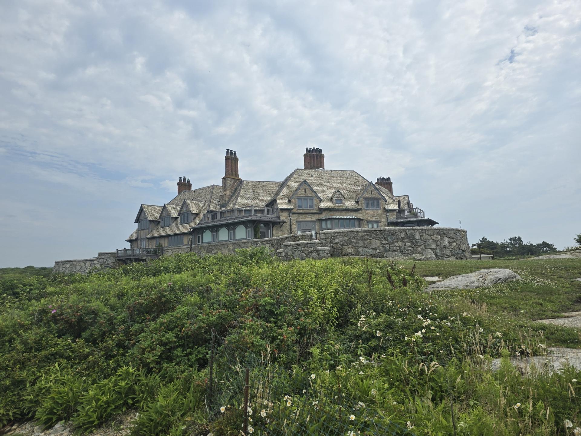 Large house on a green, overgrown hill under a cloudy sky.