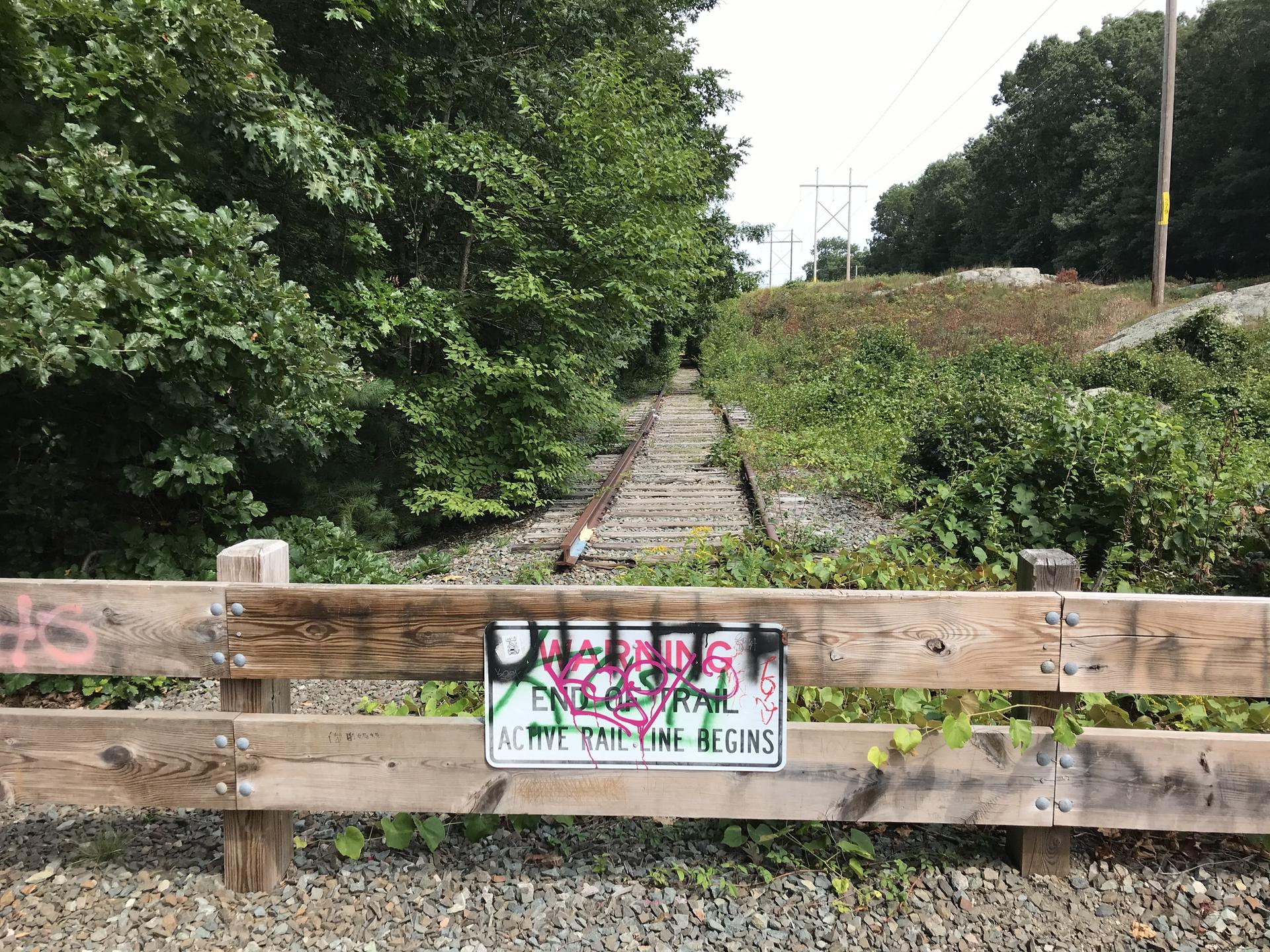 Warning sign on a wooden fence blocking overgrown railroad tracks in daylight.