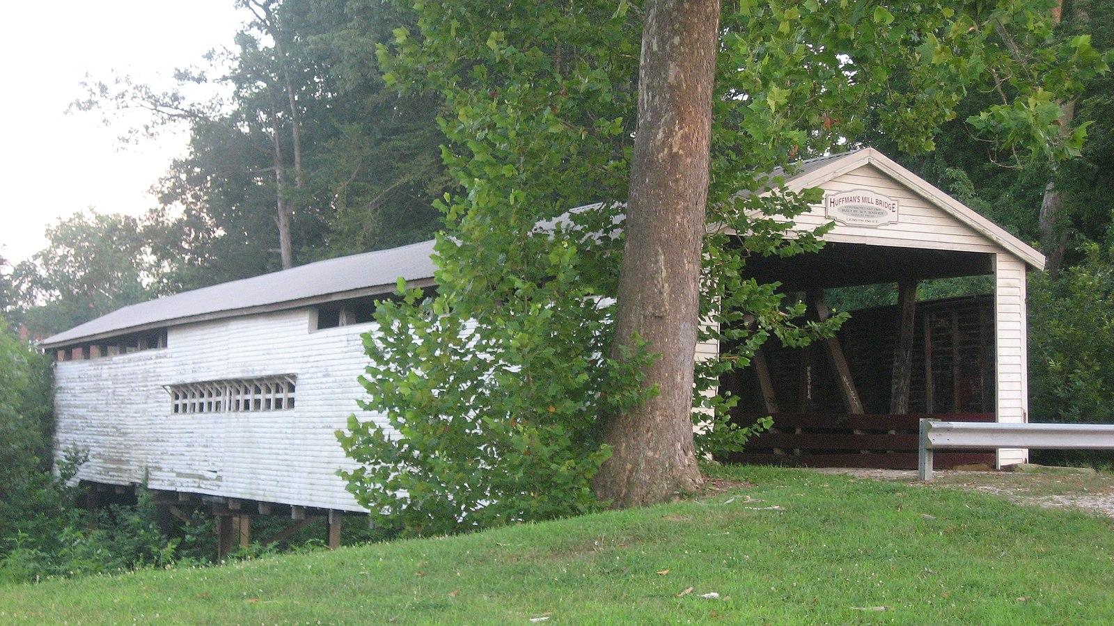 A weathered white covered bridge stands beside a large tree and green foliage.