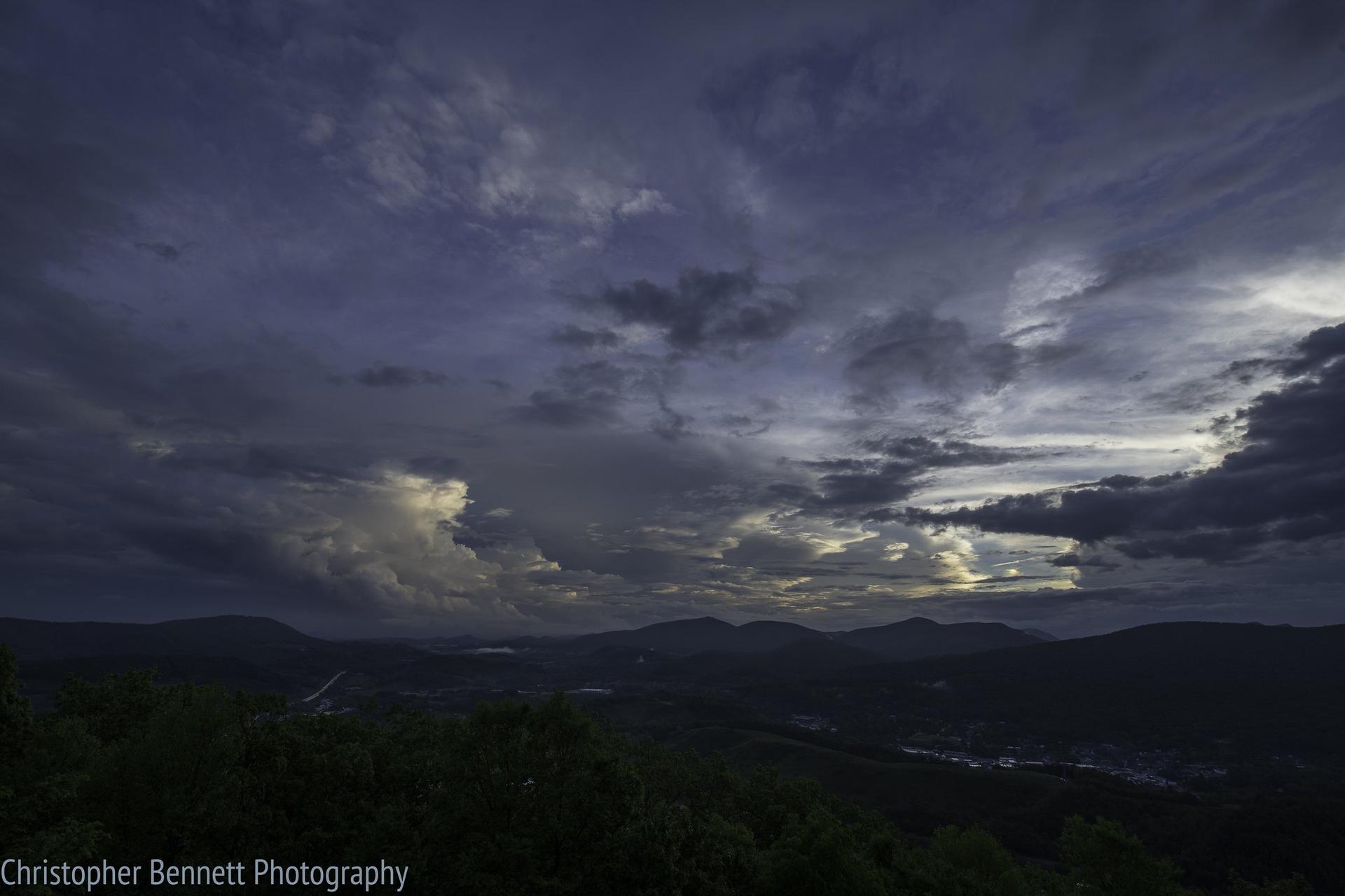 Dramatic dark clouds over a mountain landscape with distant valley lights at dusk.