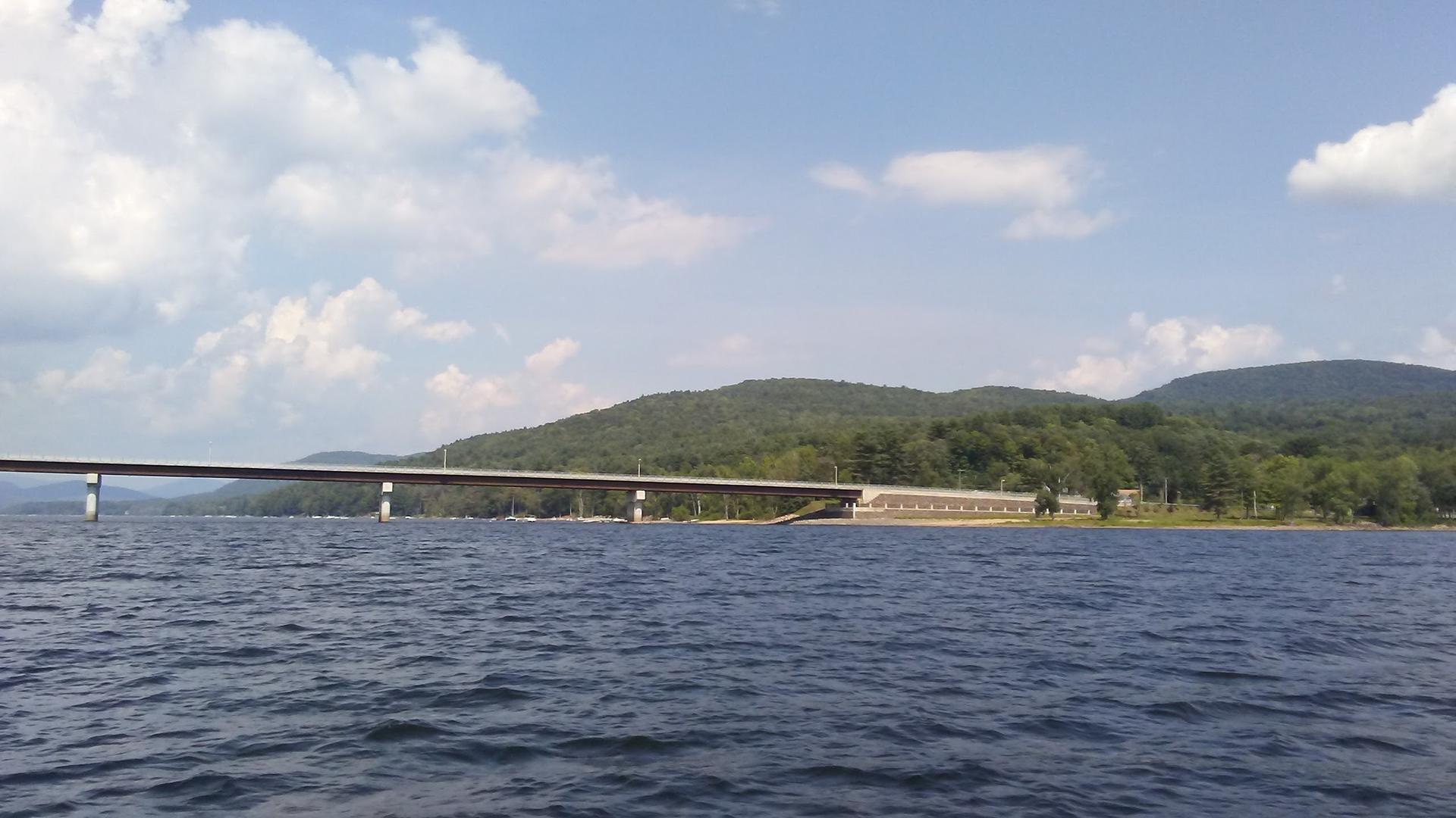 Long bridge spans dark water with forested hills under a cloudy sky.