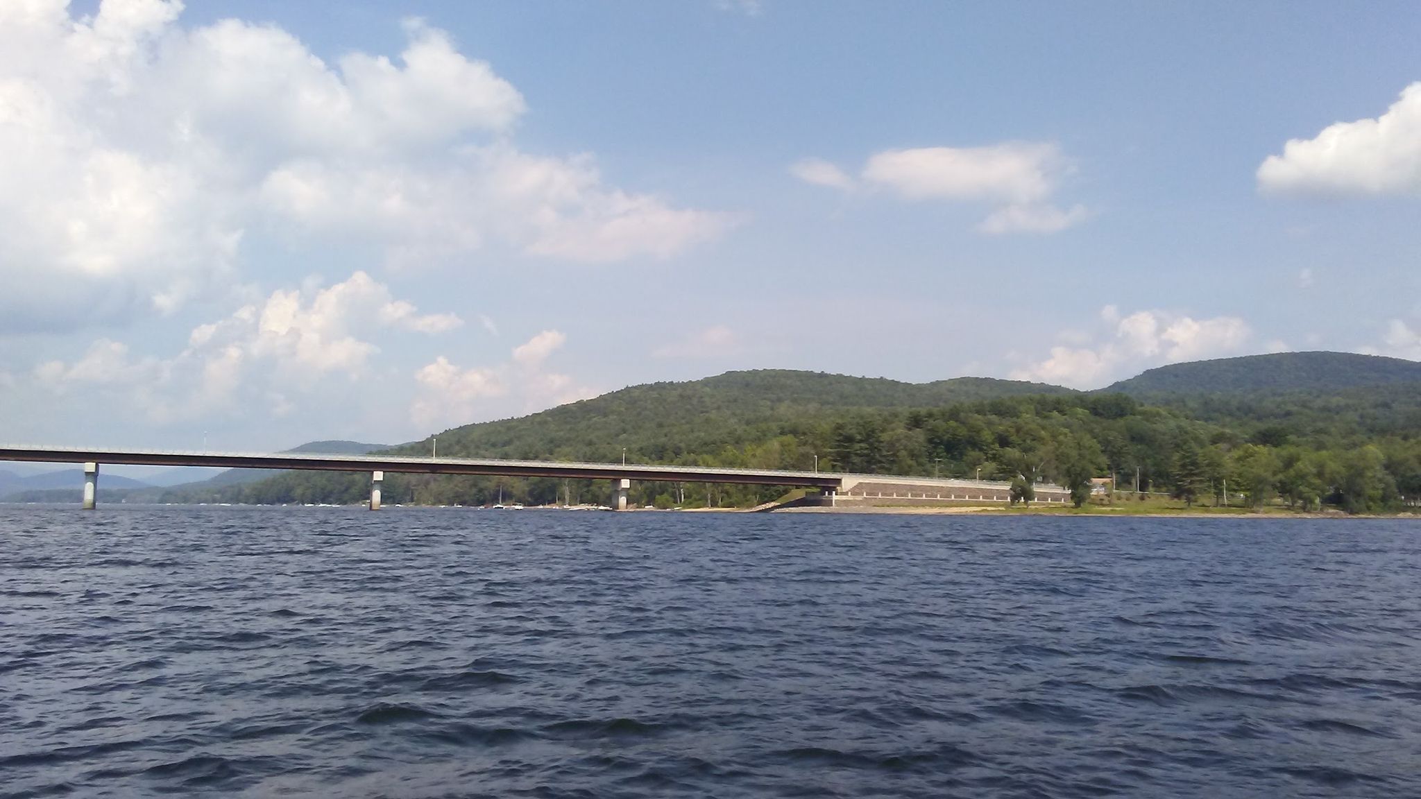 Long bridge spans dark water with forested hills under a cloudy sky. - View of Batchellerville Bridge loop from Daketown State Forest