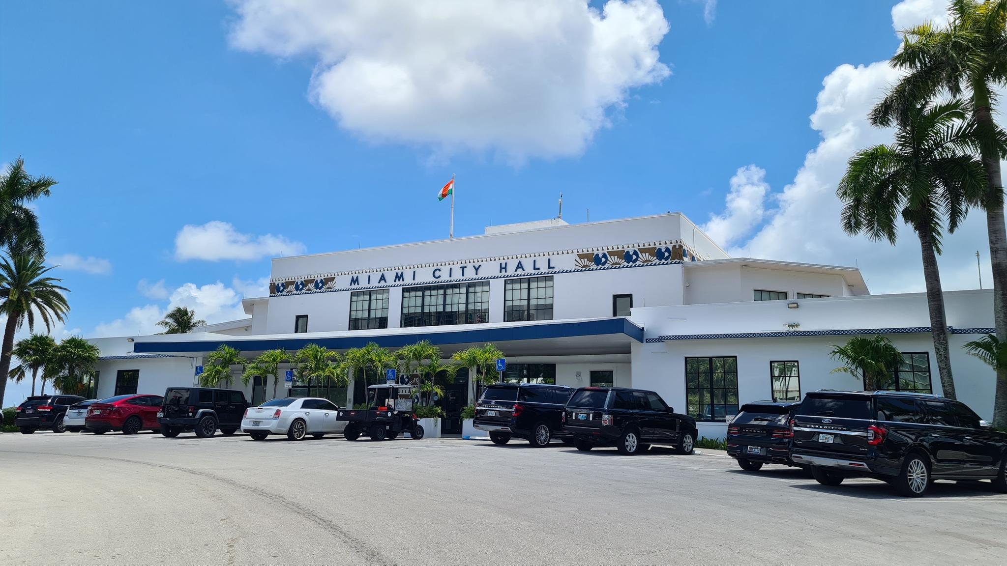 Large white building with parked cars and palm trees under a bright blue sky.