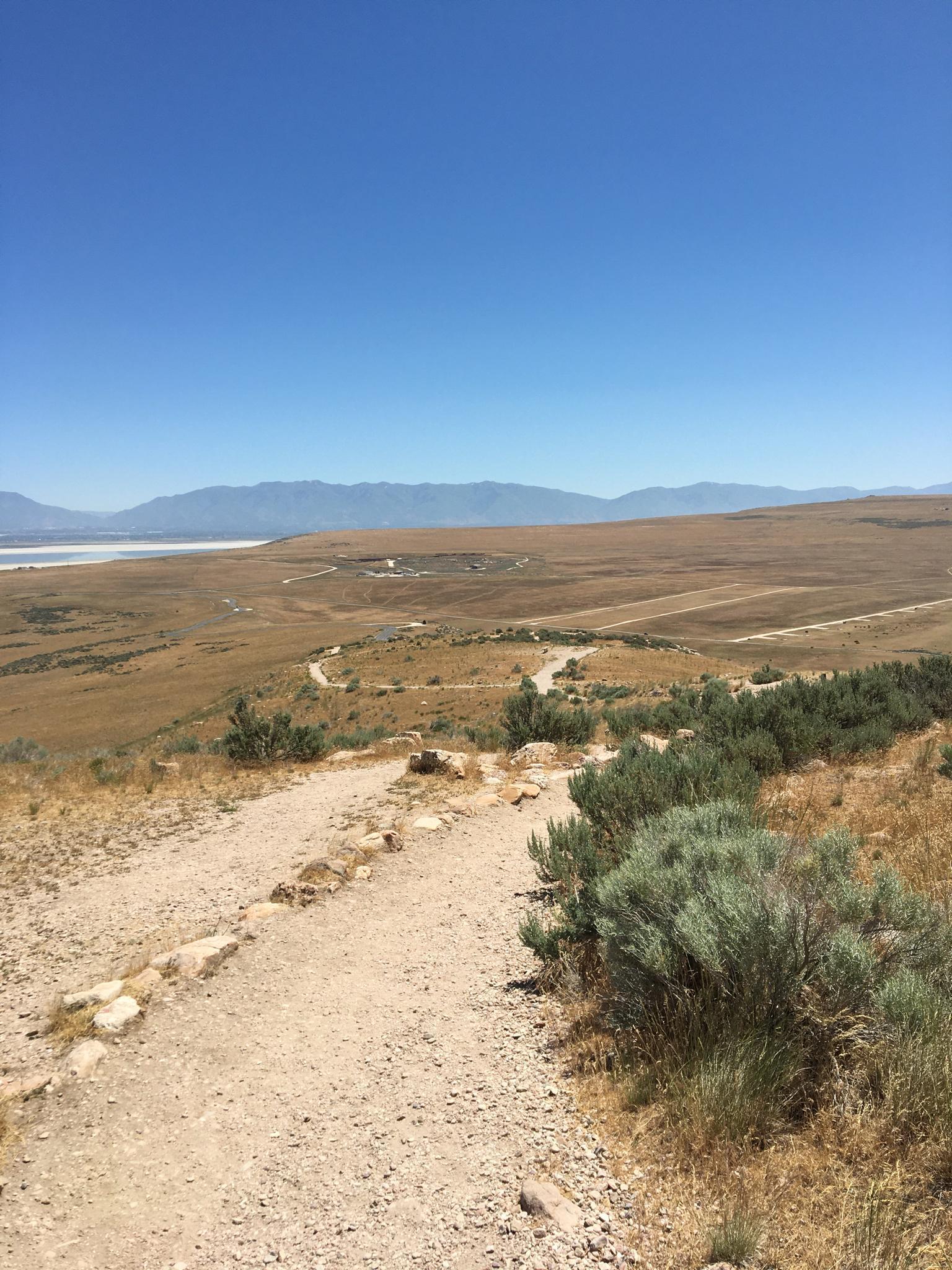 Dirt path winding through dry, grassy hills towards a distant lake and mountains.
