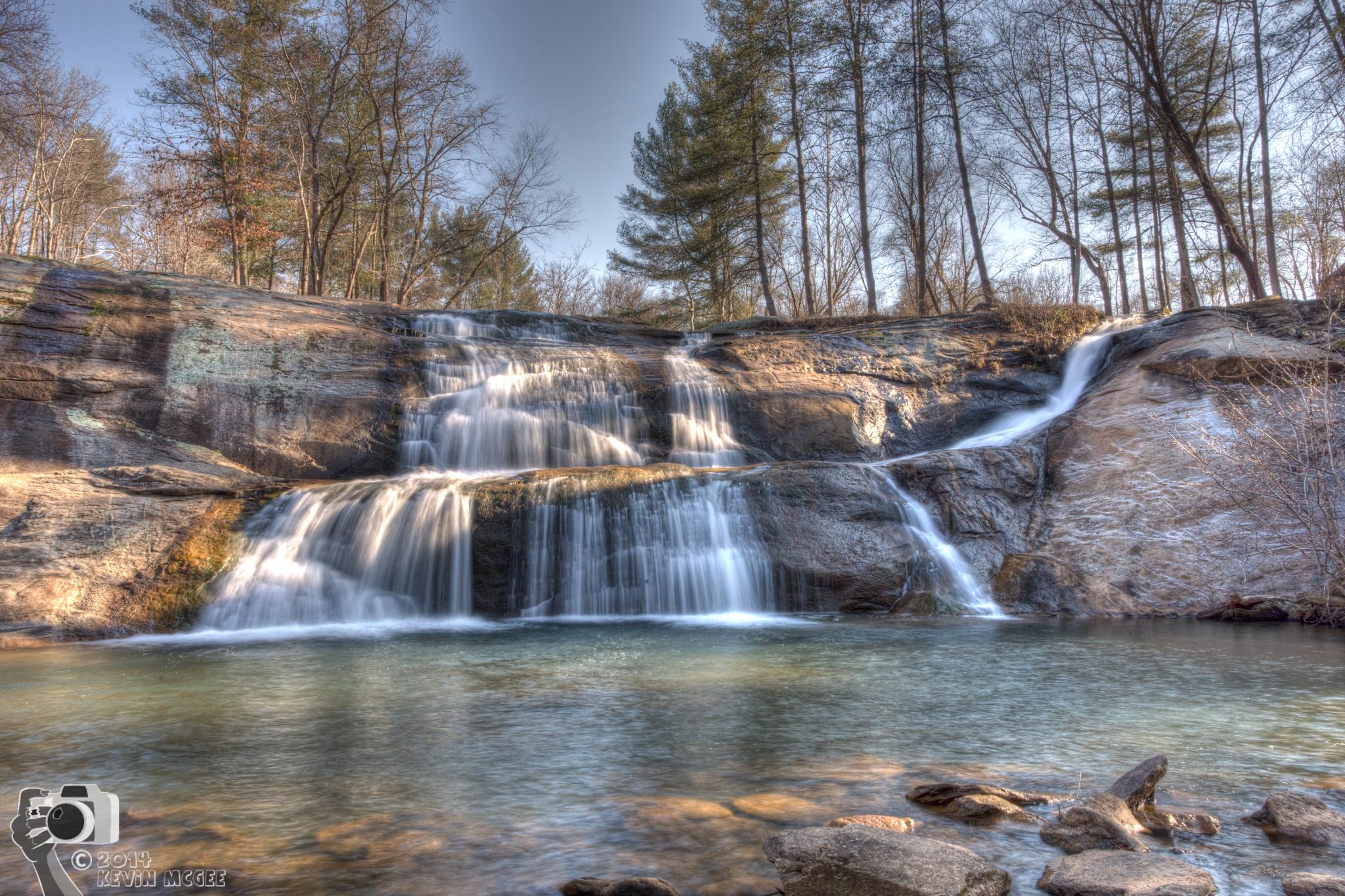 Multi-tiered waterfall cascades into a clear pool under bright sunlight.