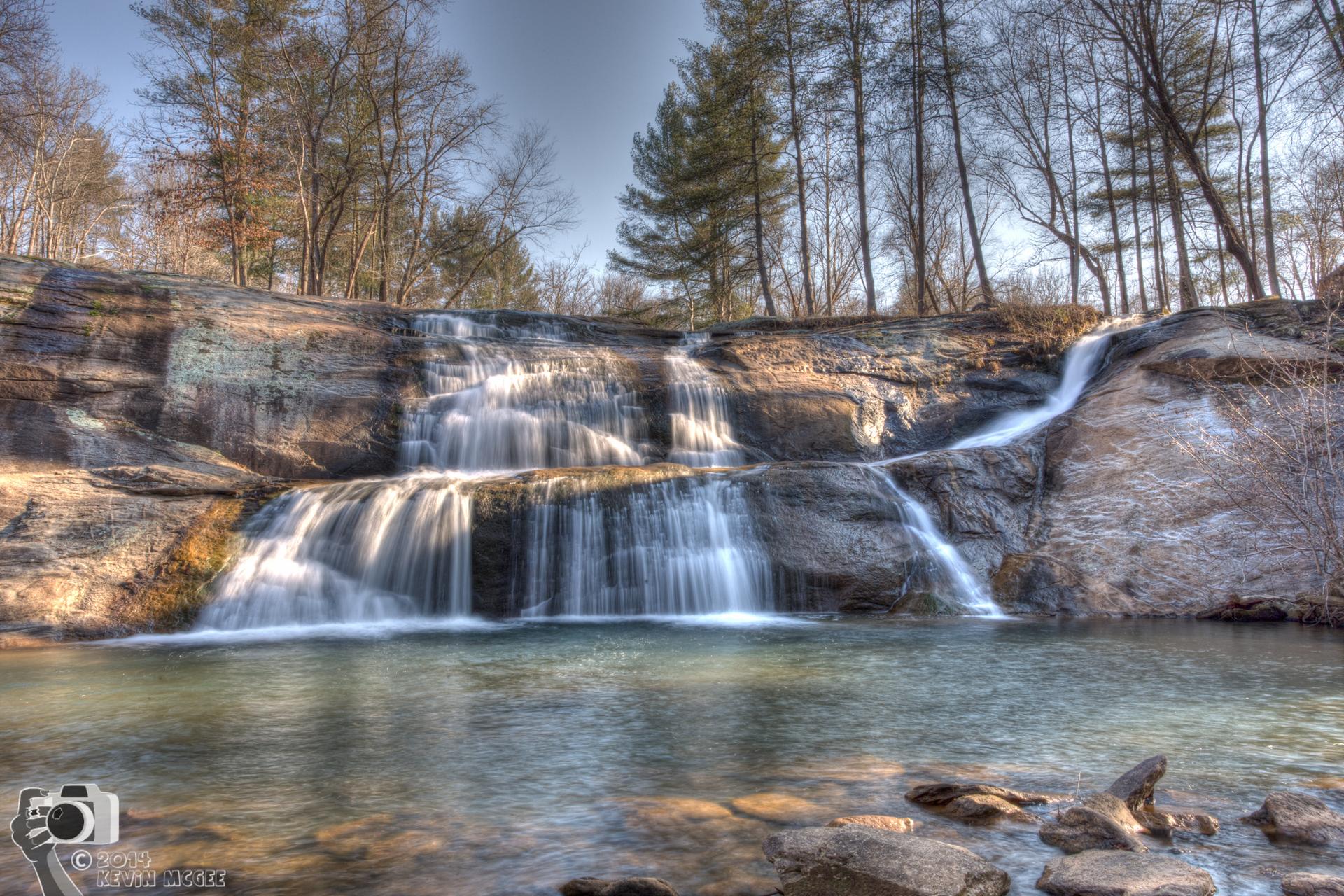 Multi-tiered waterfall cascades into a clear pool under bright sunlight.