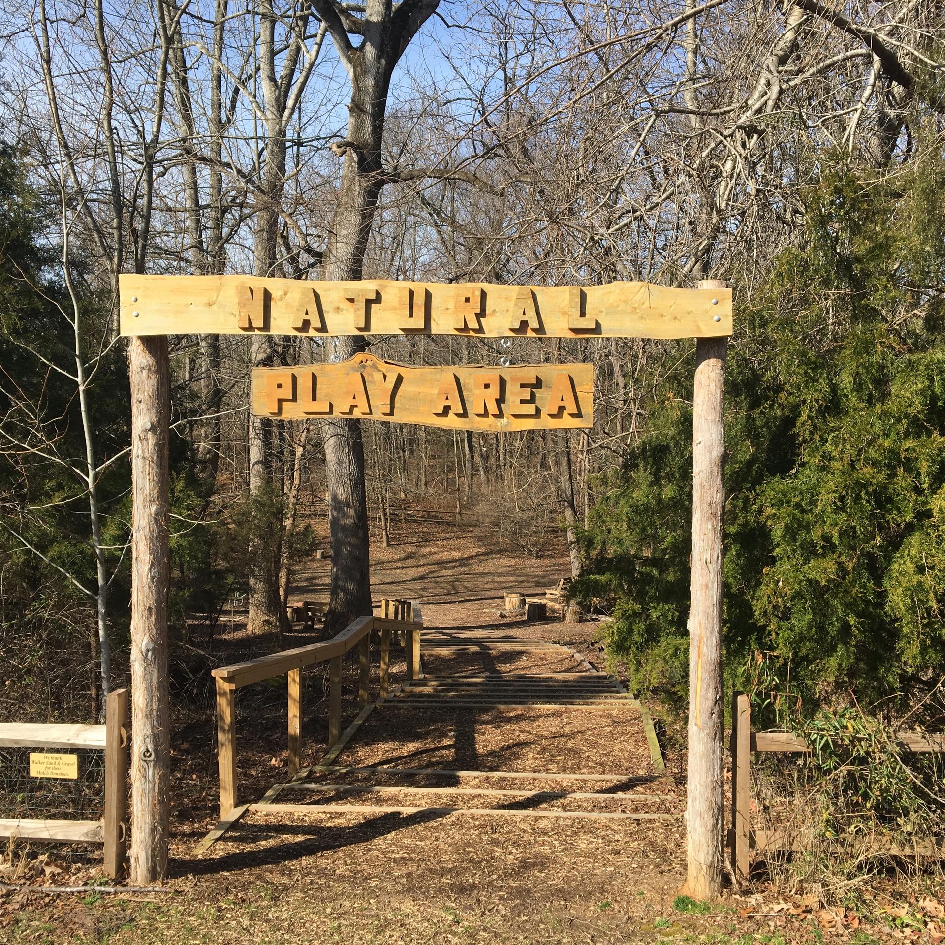 Wooden sign 'NATURAL PLAY AREA' over a sunny path leading into bare woods.
