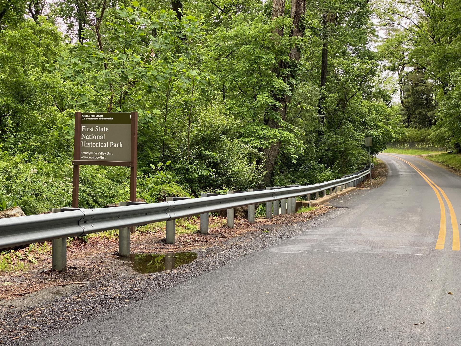 A paved road with a guardrail next to a sign and green trees.