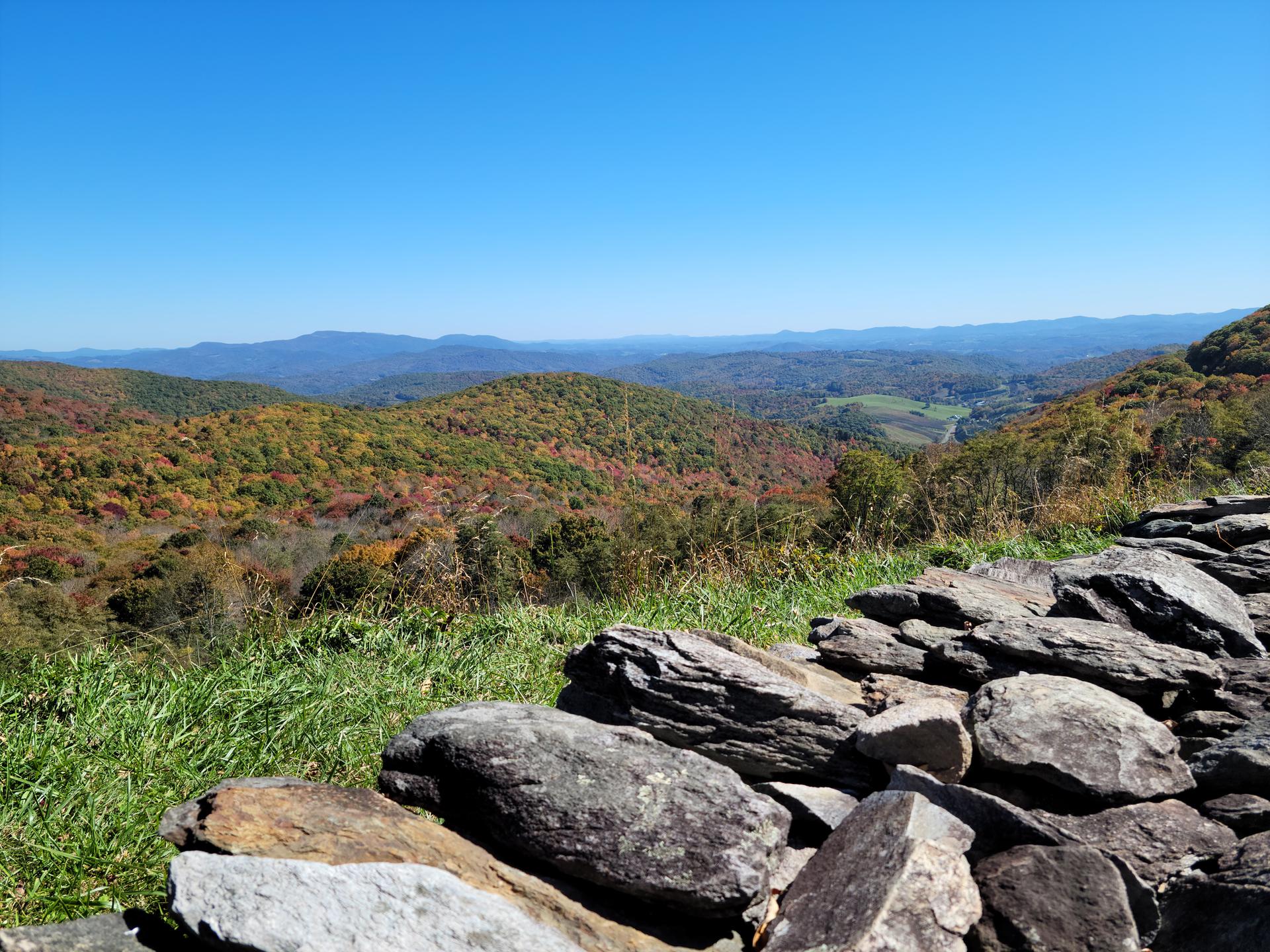 Sunny mountain vista with autumn trees, distant peaks, and a rocky foreground.