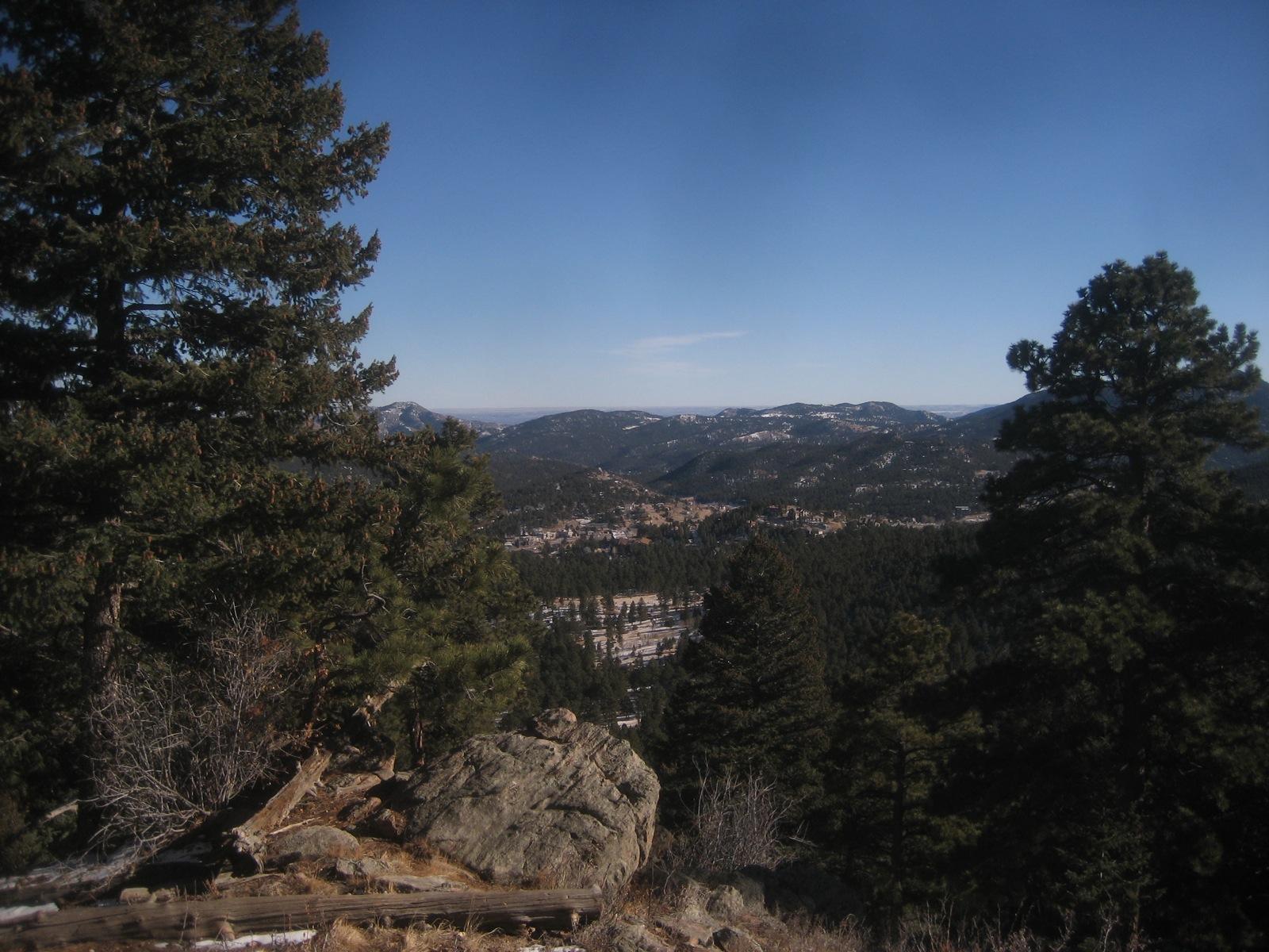 Evergreen trees frame a vast valley and snow-dusted mountains on a clear day.