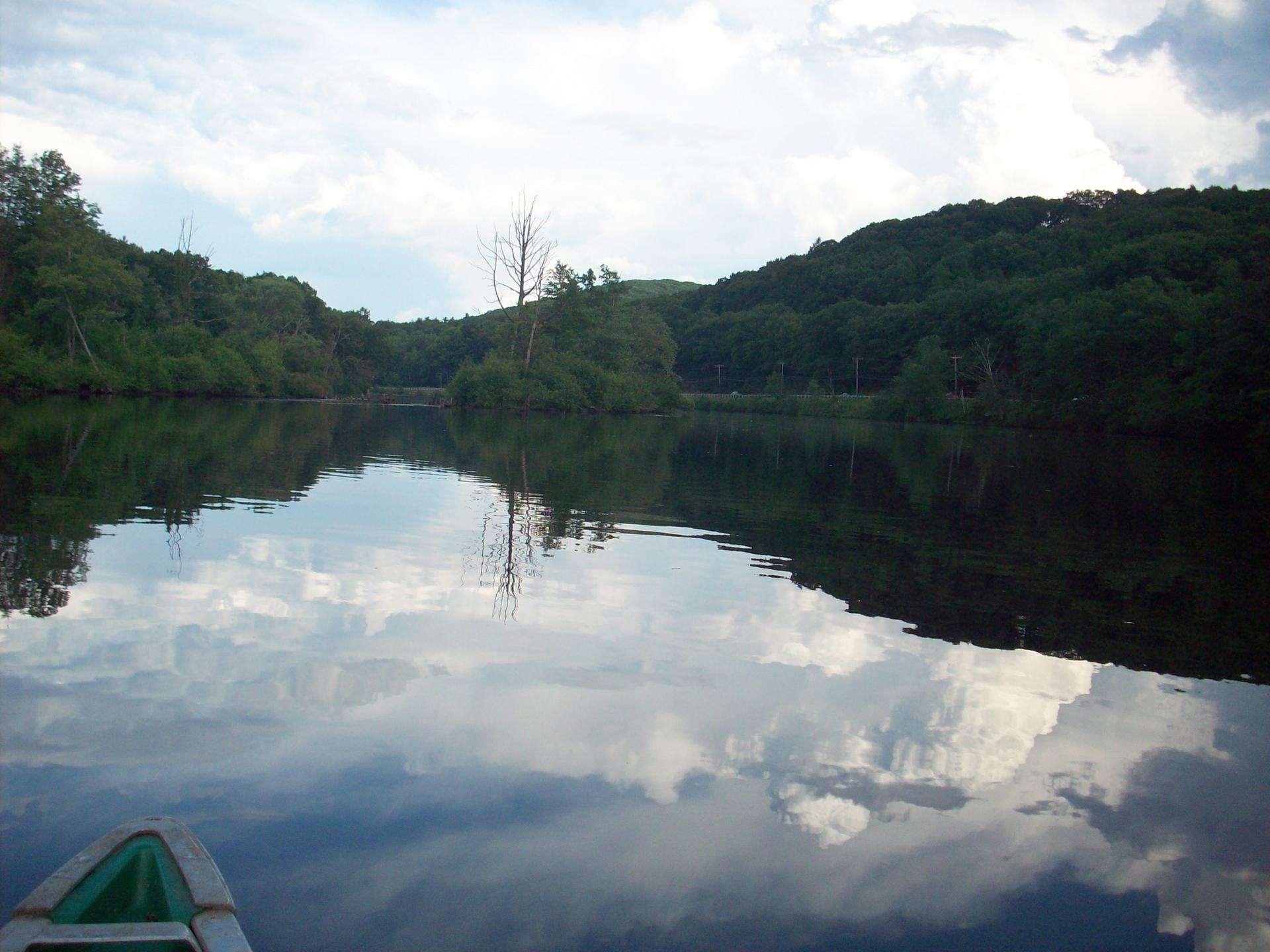 Calm water reflecting cloudy sky, surrounded by forested hills, with a boat in foreground.