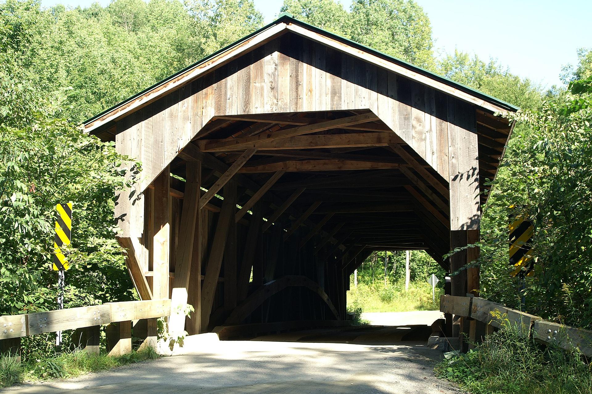 A rustic wooden covered bridge on a sunny day, surrounded by green trees.