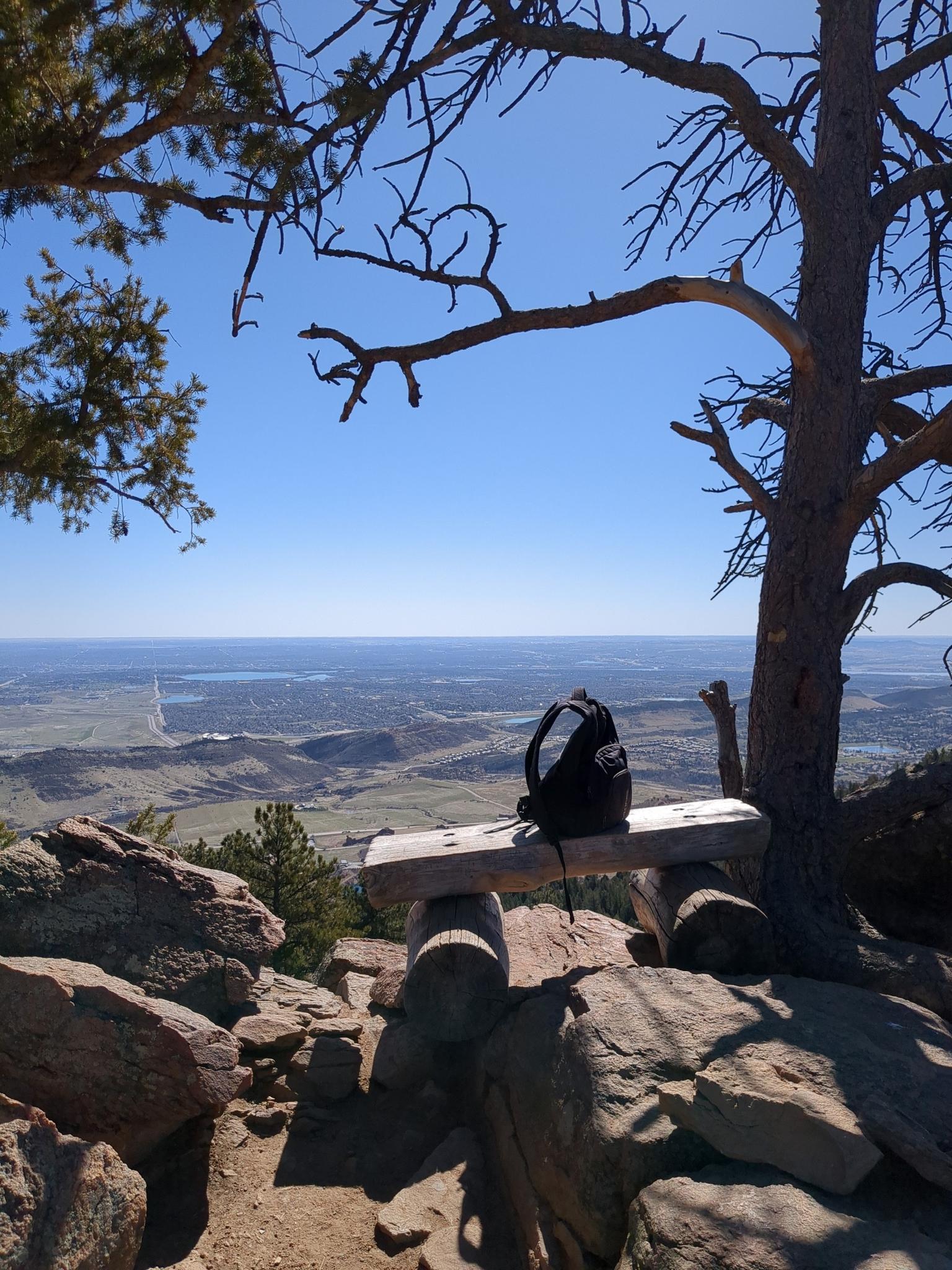 Backpack on a wooden bench overlooking a vast sunny valley from a high vantage.