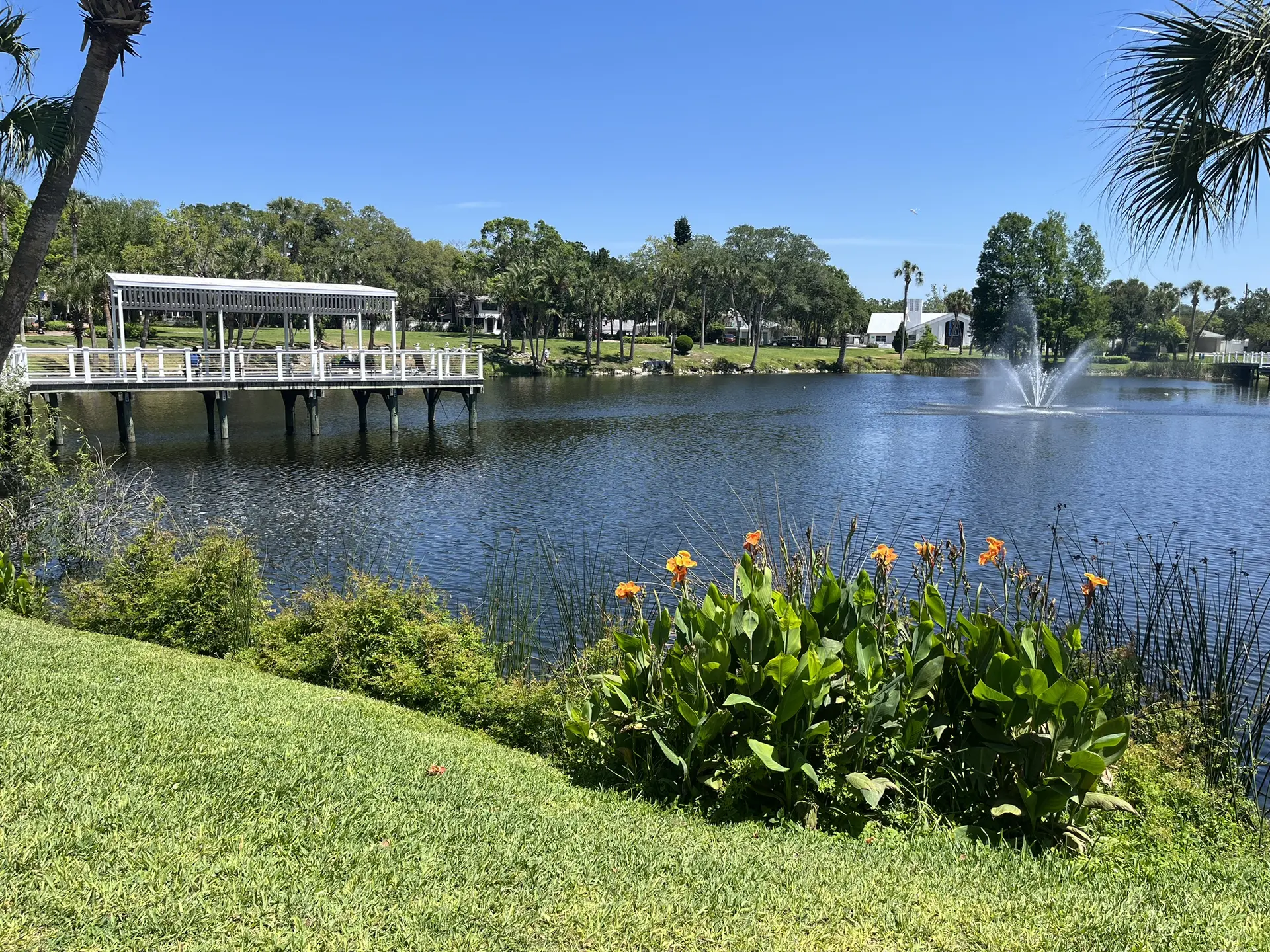 Sunny lake with a fountain, white gazebo, and vibrant green foliage.