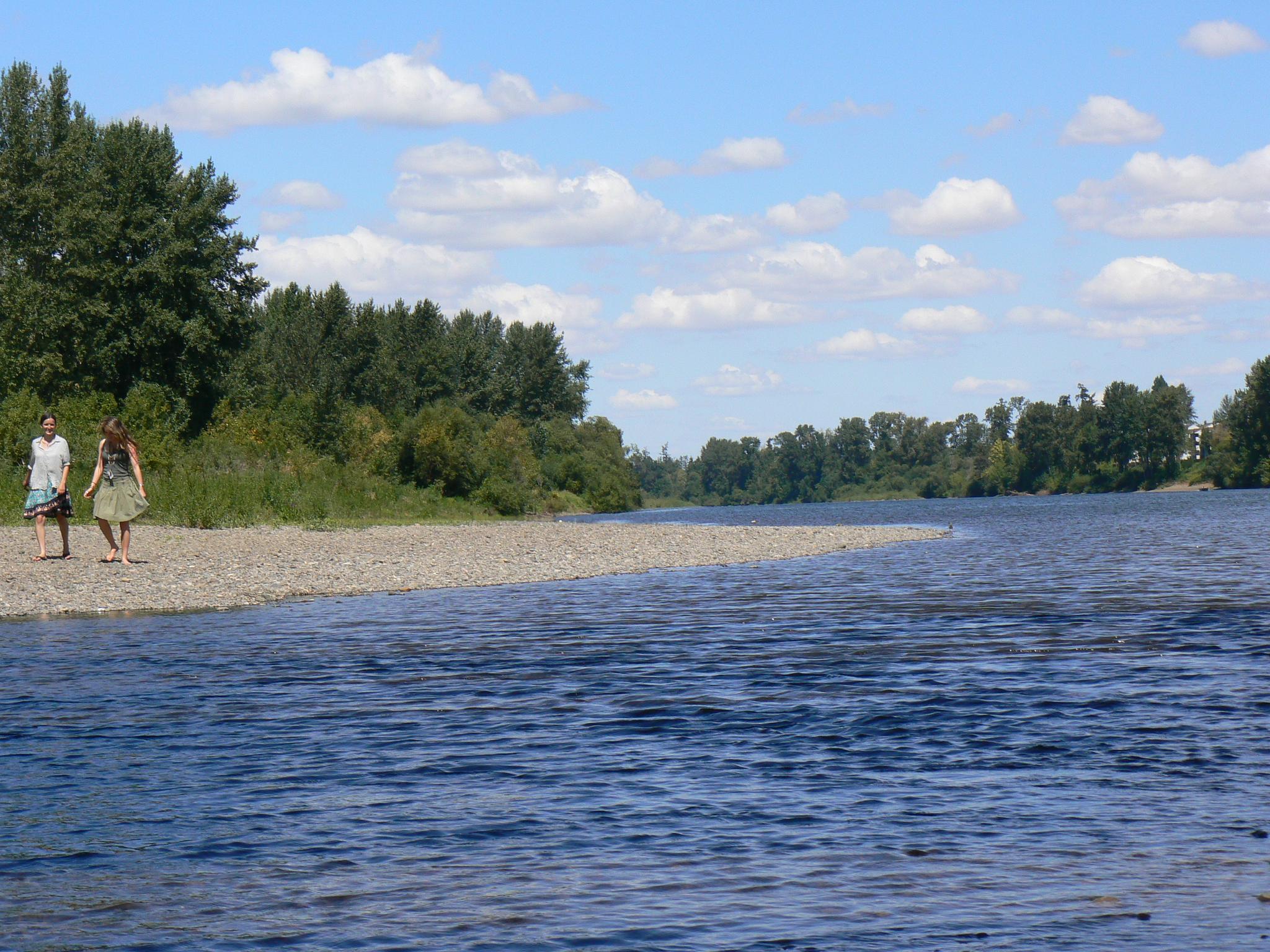 Two people stand on a sunny gravel riverbank next to a wide river.