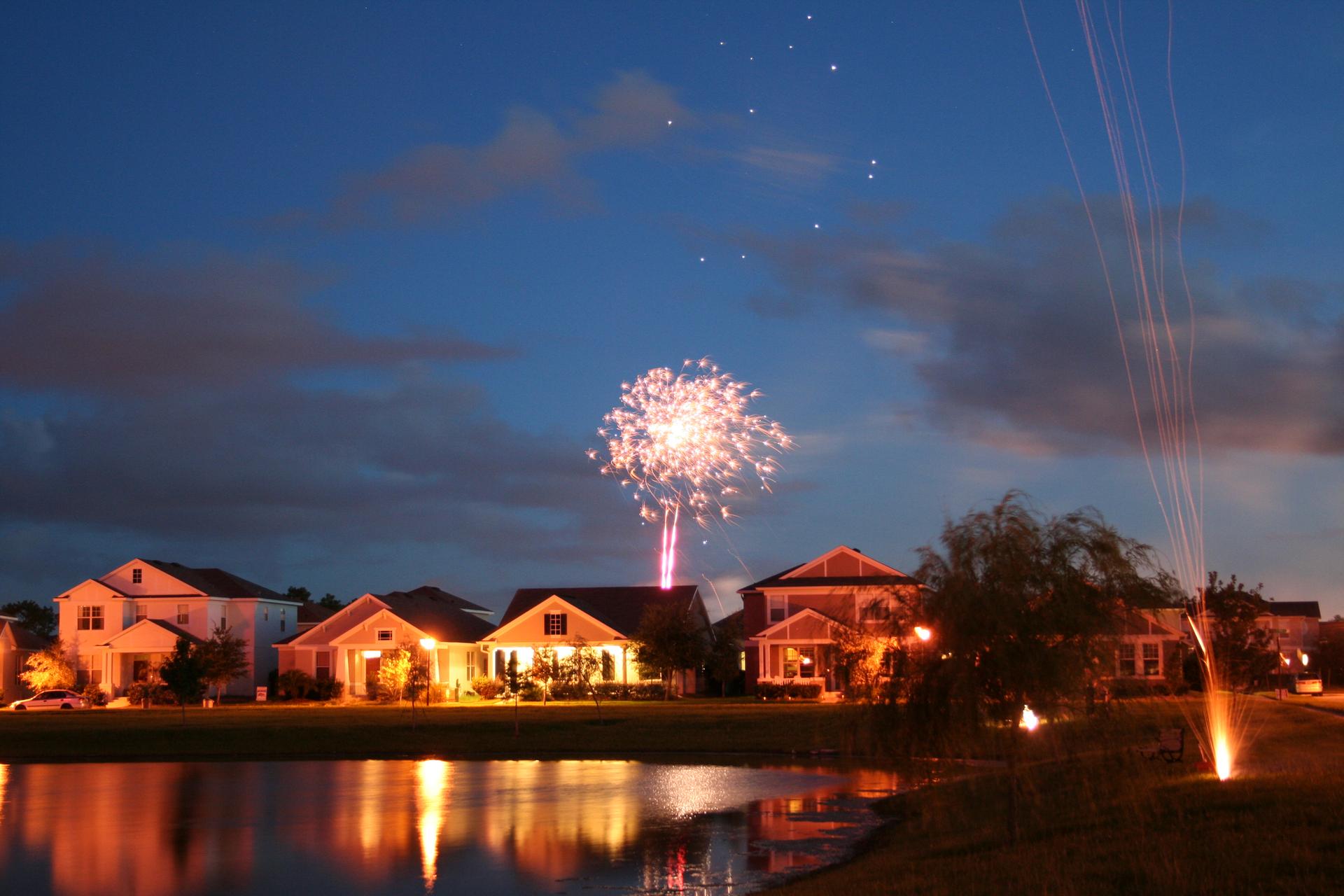 Fireworks explode over houses and a lake at night, reflecting on the water.