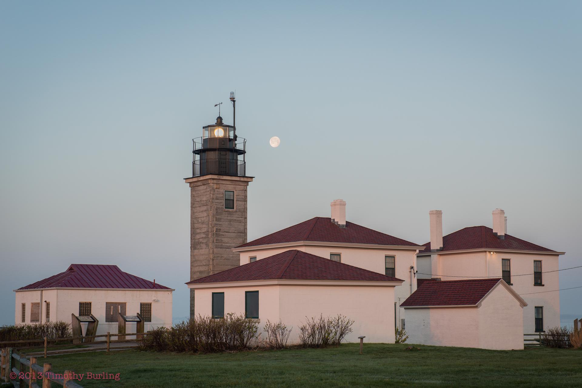 Lighthouse and white buildings under a soft evening sky with a full moon.
