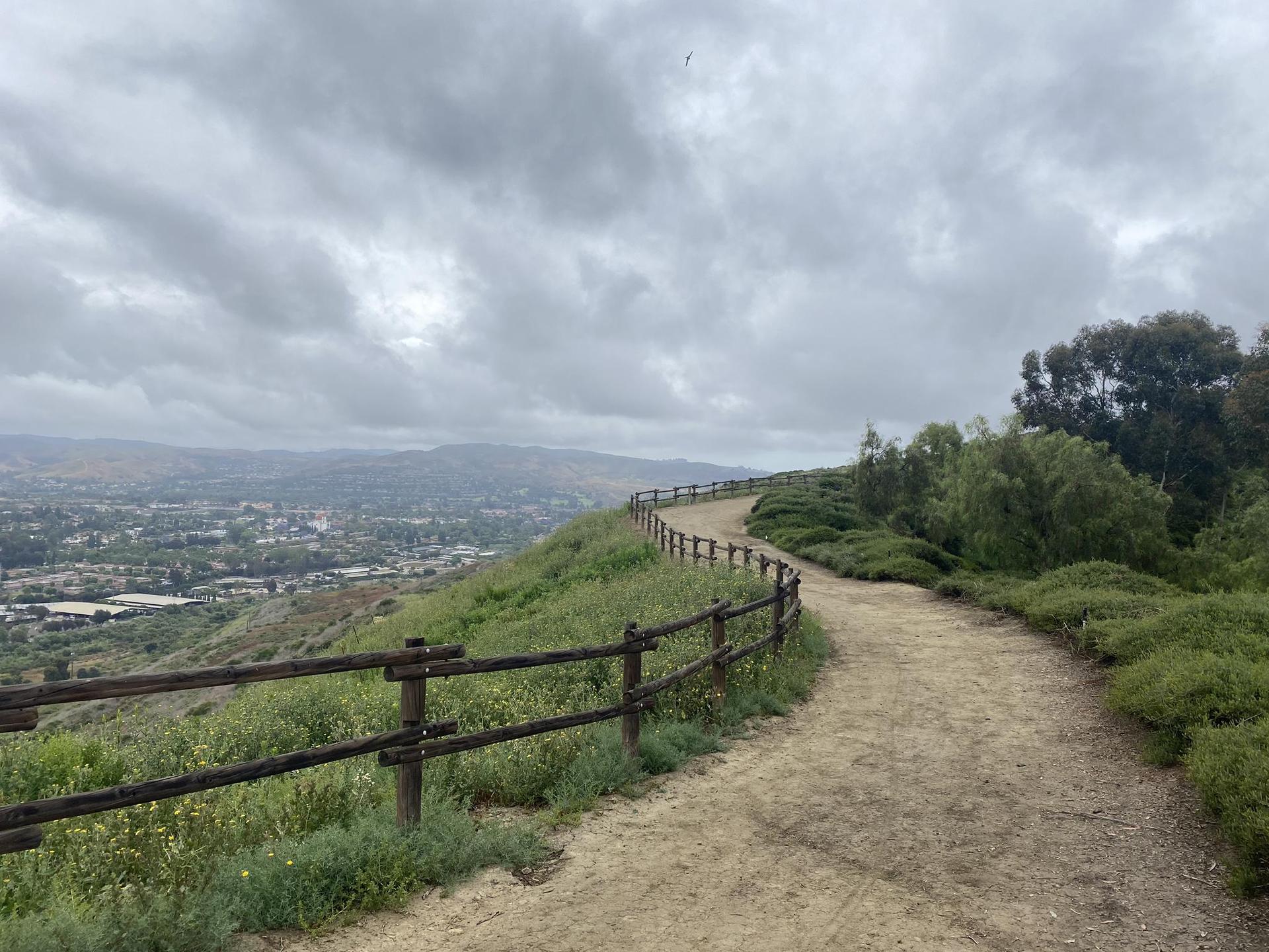 Winding dirt path with a wooden fence overlooking a city on an overcast day.