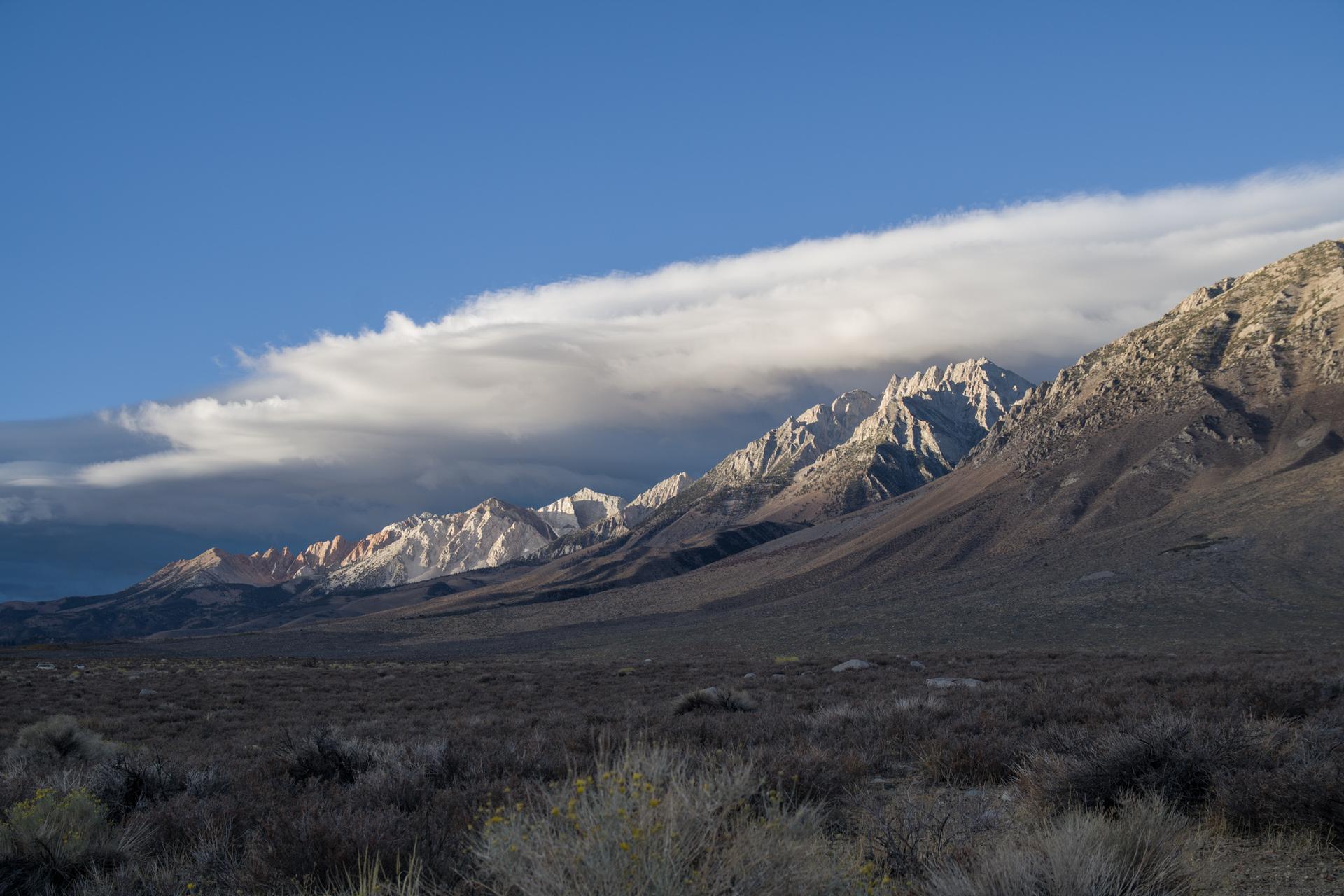 Dramatic clouds partially obscure rugged mountains and arid foreground under bright sun.