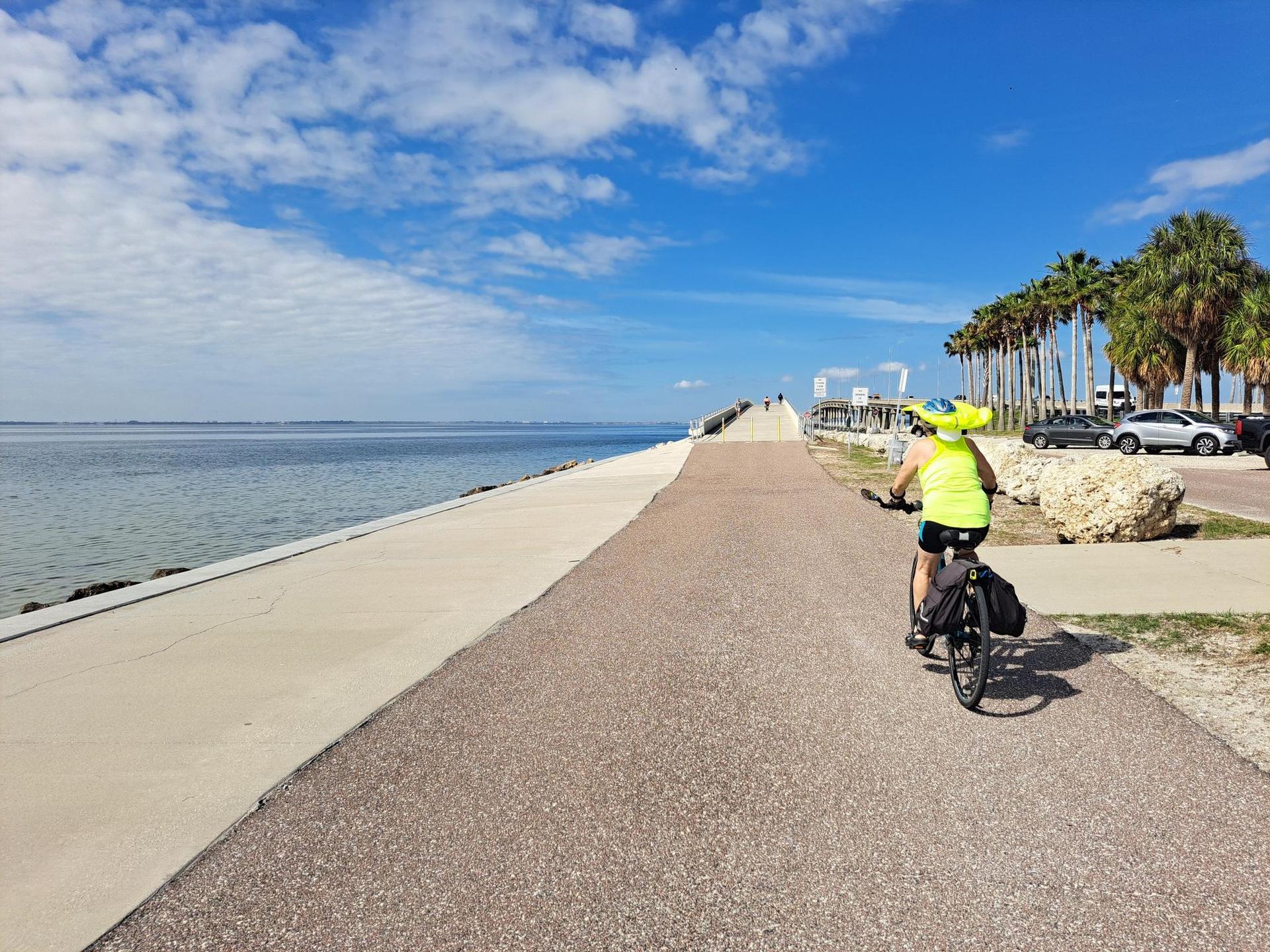 Person cycling on a paved path next to calm water under a bright blue sky.