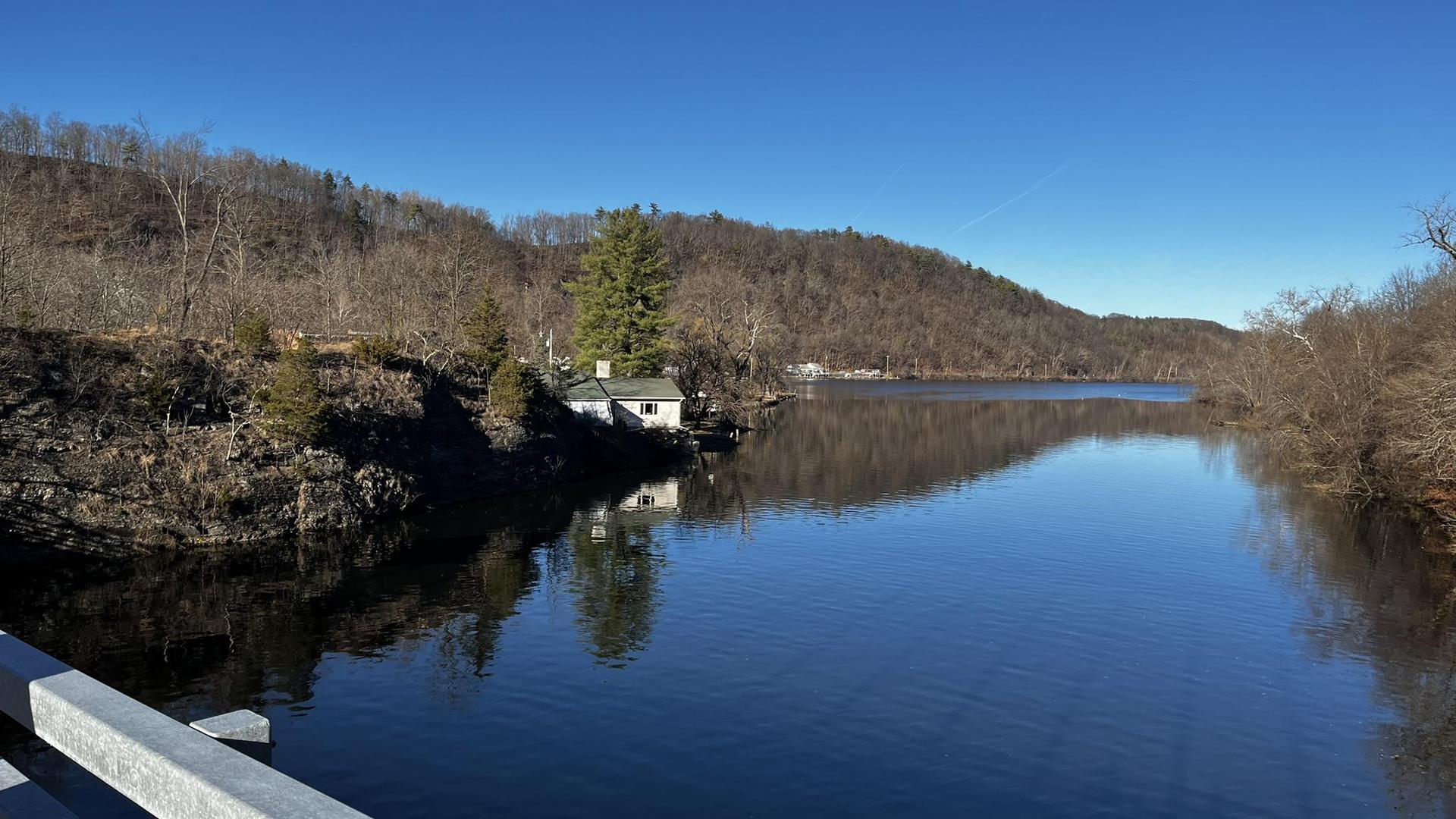Calm river reflecting wooded hills and a small house under a clear blue sky.