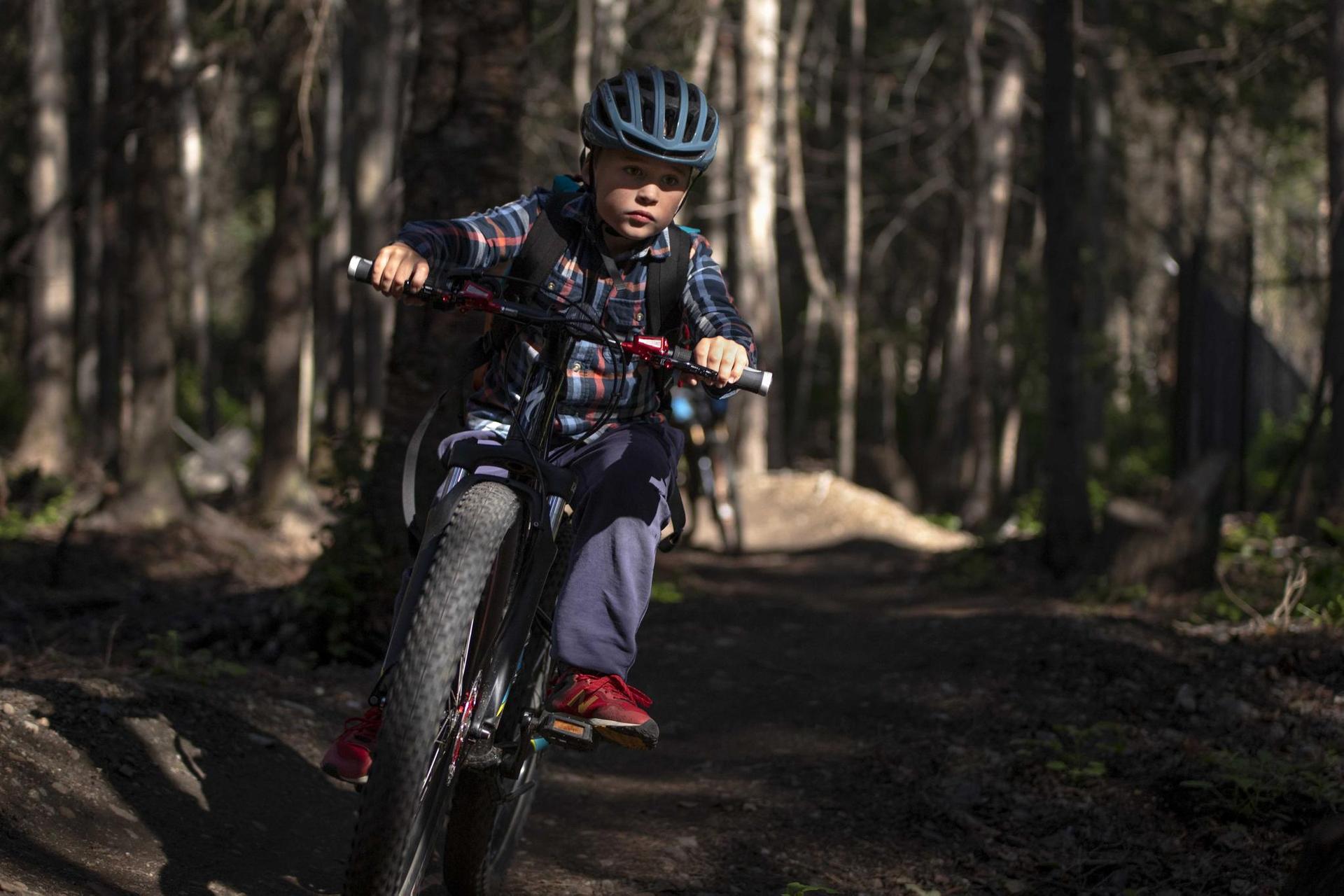 Young person in helmet and backpack mountain biking on a sun-dappled forest trail.