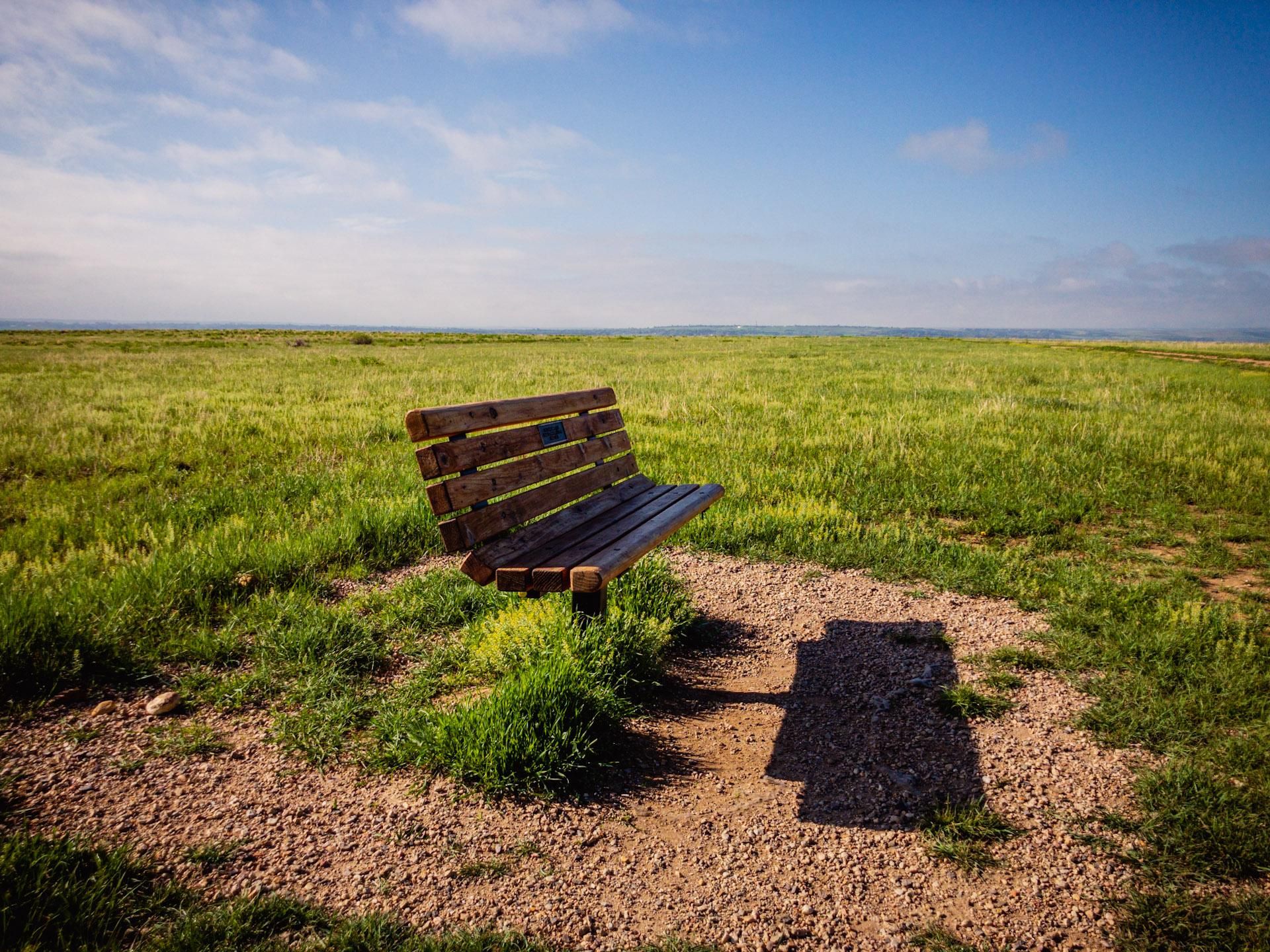 A wooden bench sits on a gravel patch in a vast sunny green field. - Gunbarrel East Boulder Trail Loop