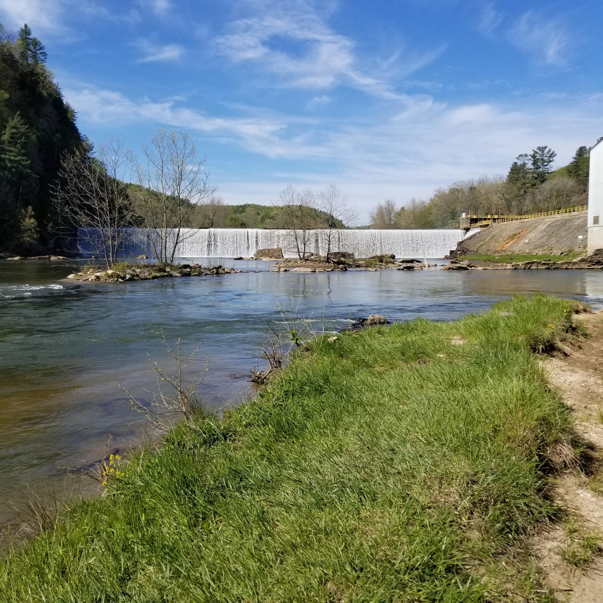 Wide river flowing towards a dam, with green grass and forested hills.