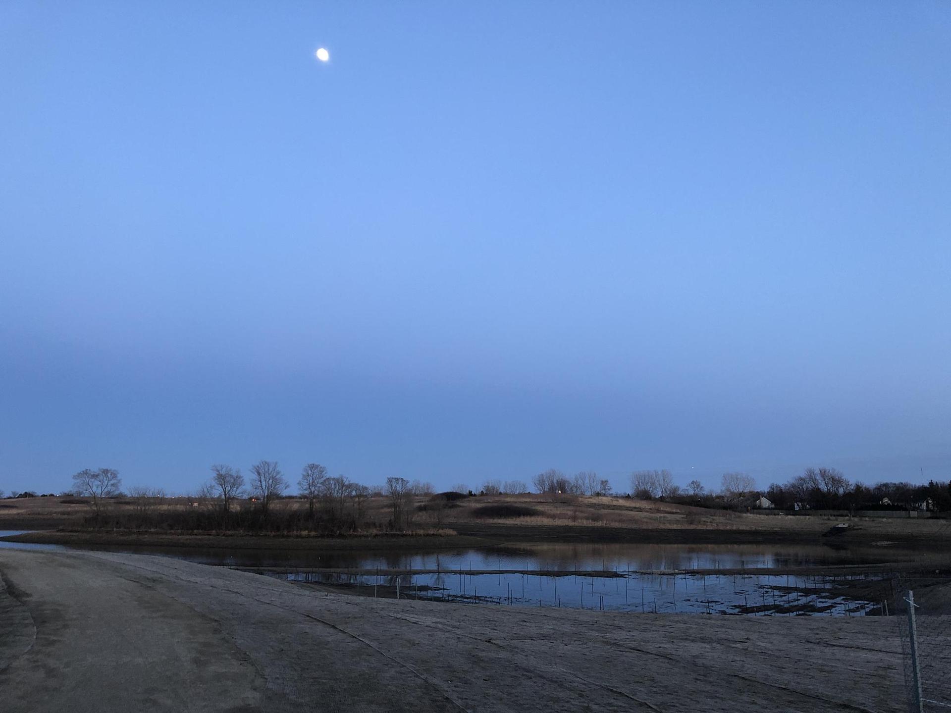Moon in blue twilight sky over calm water and distant land.