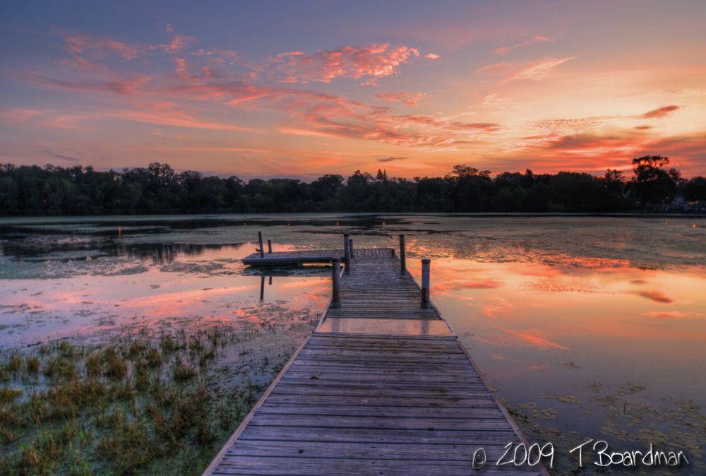 Wooden dock extending into a calm lake reflecting a vibrant orange and pink sunset.