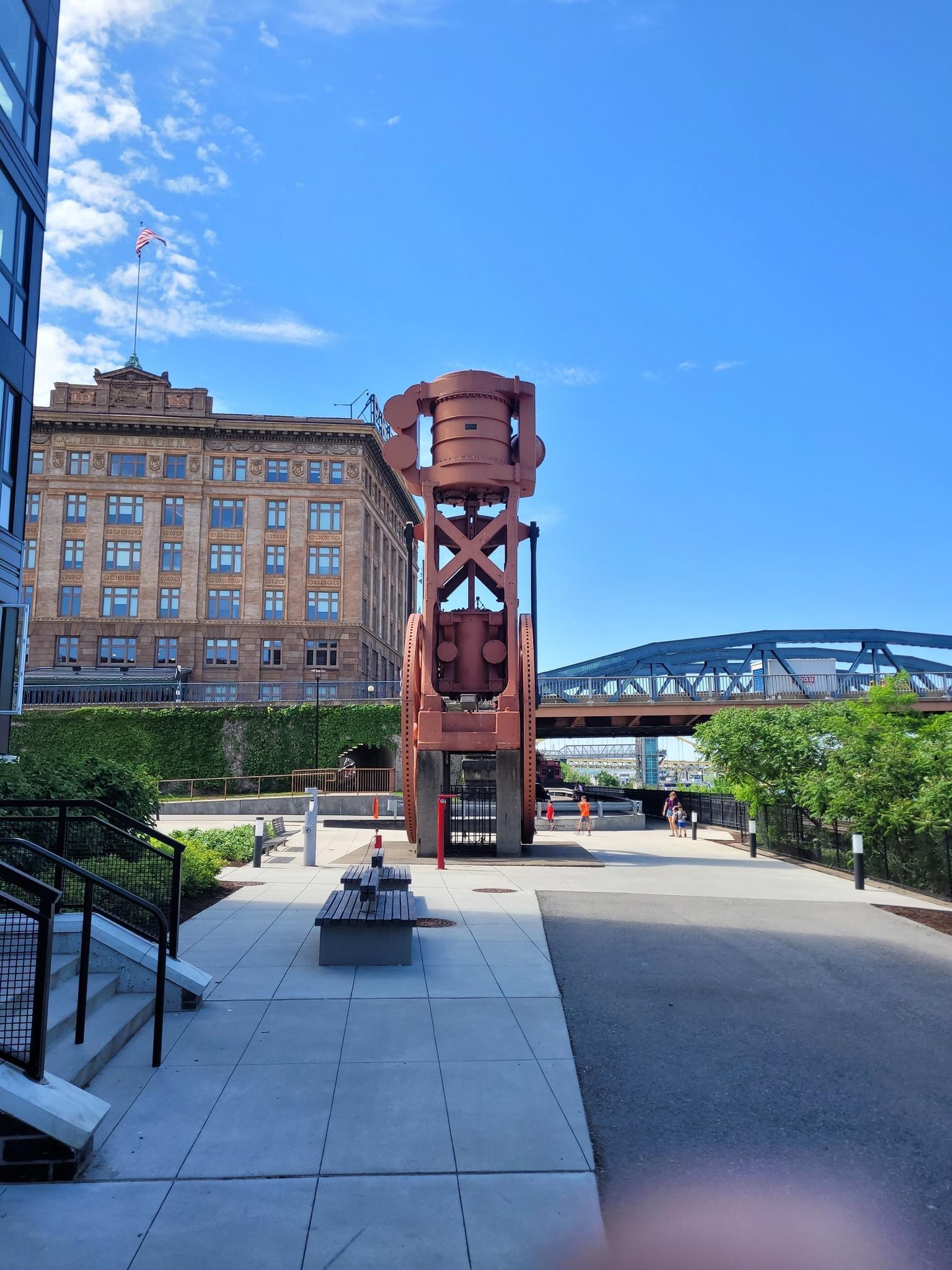 A large red industrial sculpture stands on a sunny urban walkway.