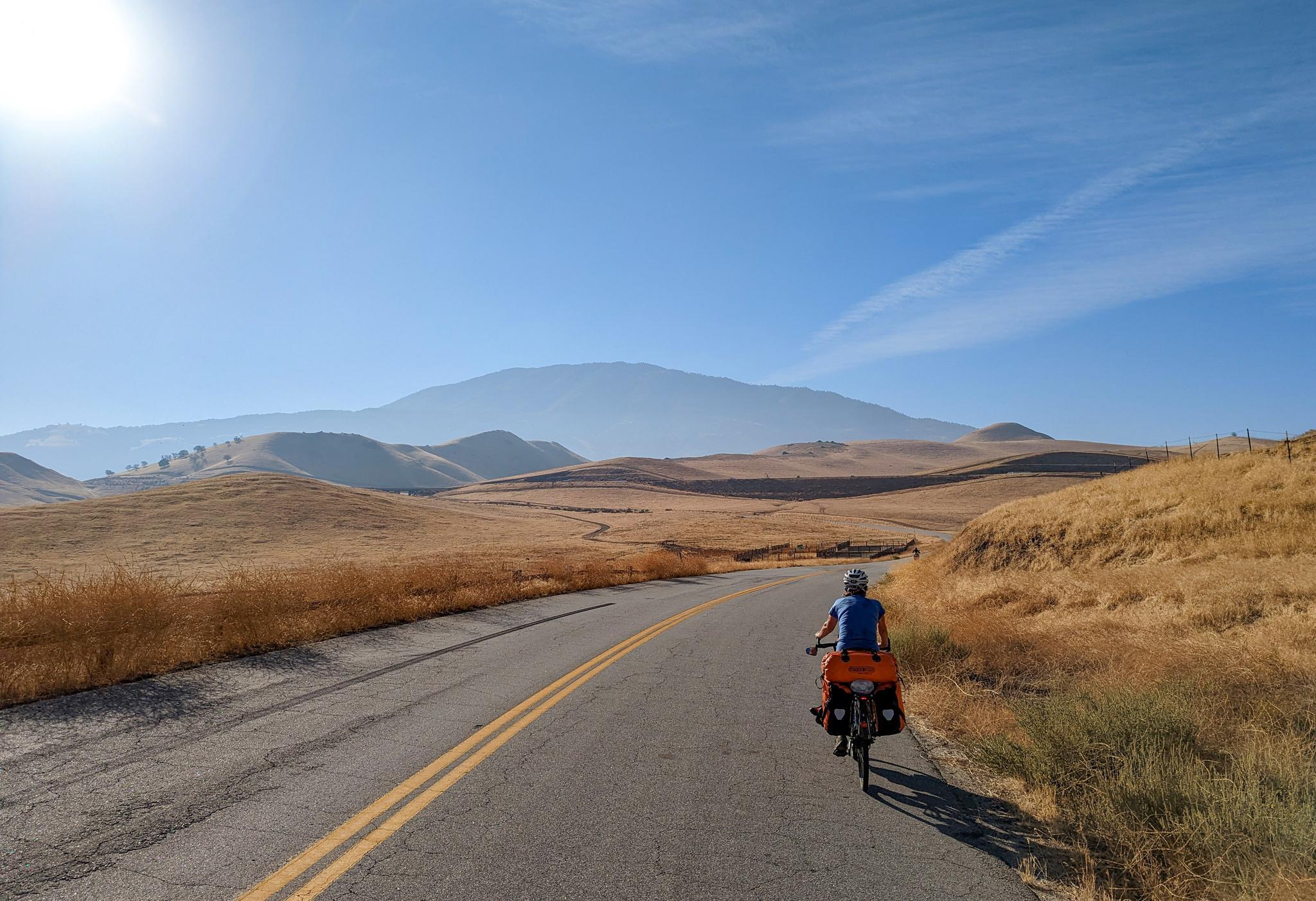 Cyclist rides on a paved road through dry, rolling hills under a bright sun.