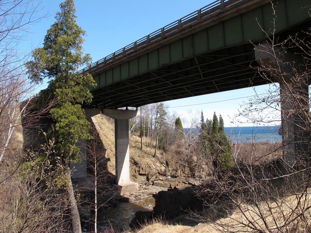 Large bridge spans a rocky gorge with trees and distant water on a sunny day.