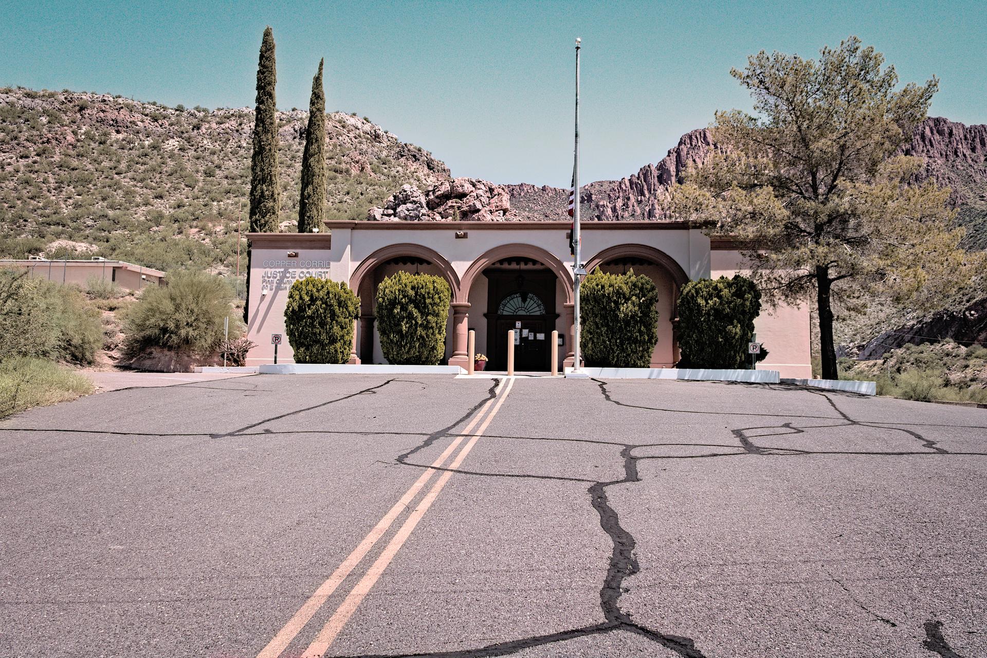 Arched building with tall trees and flagpole, desert mountains under bright sky.
