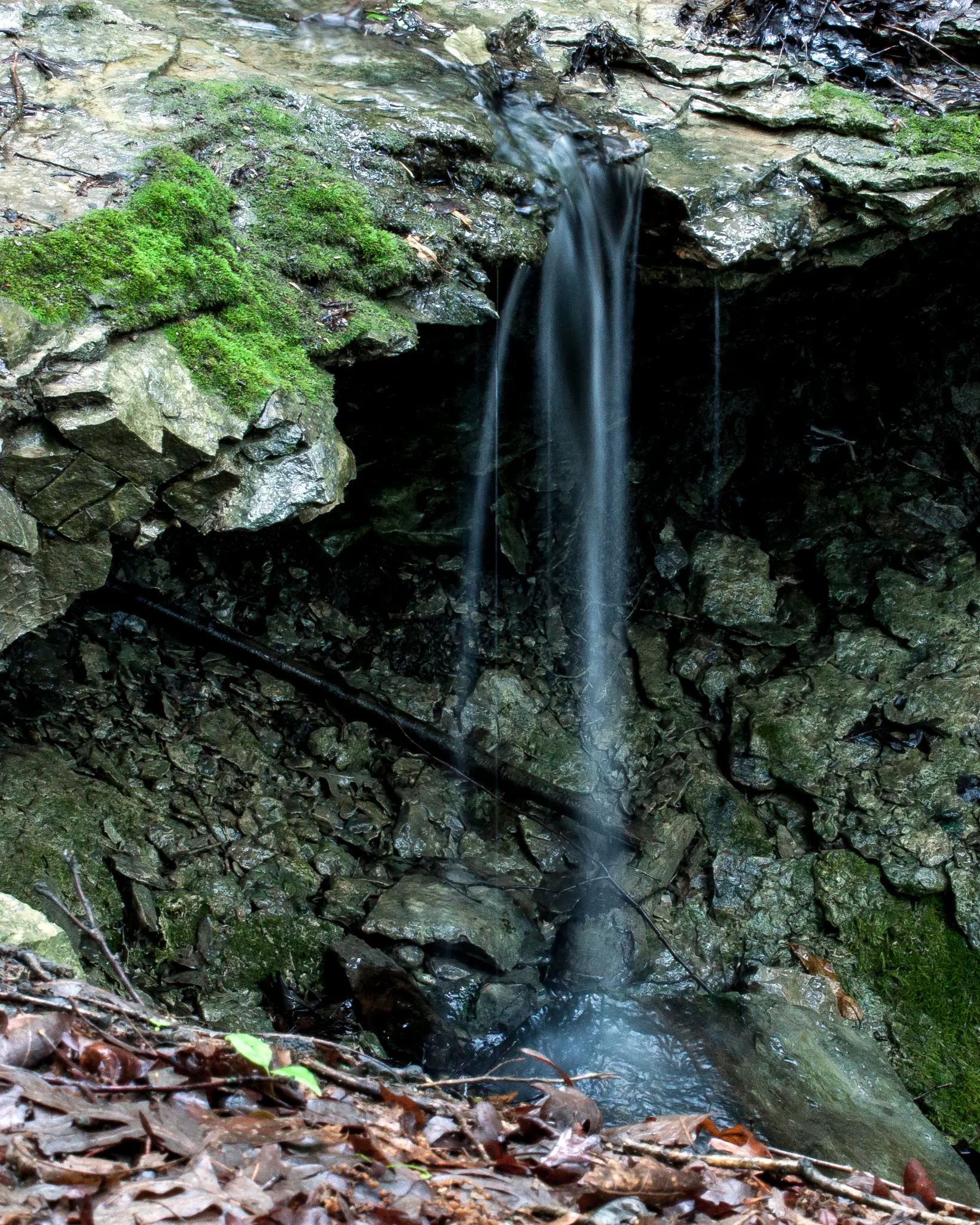 A small, blurry waterfall cascades down moss-covered rocks into a dark pool.