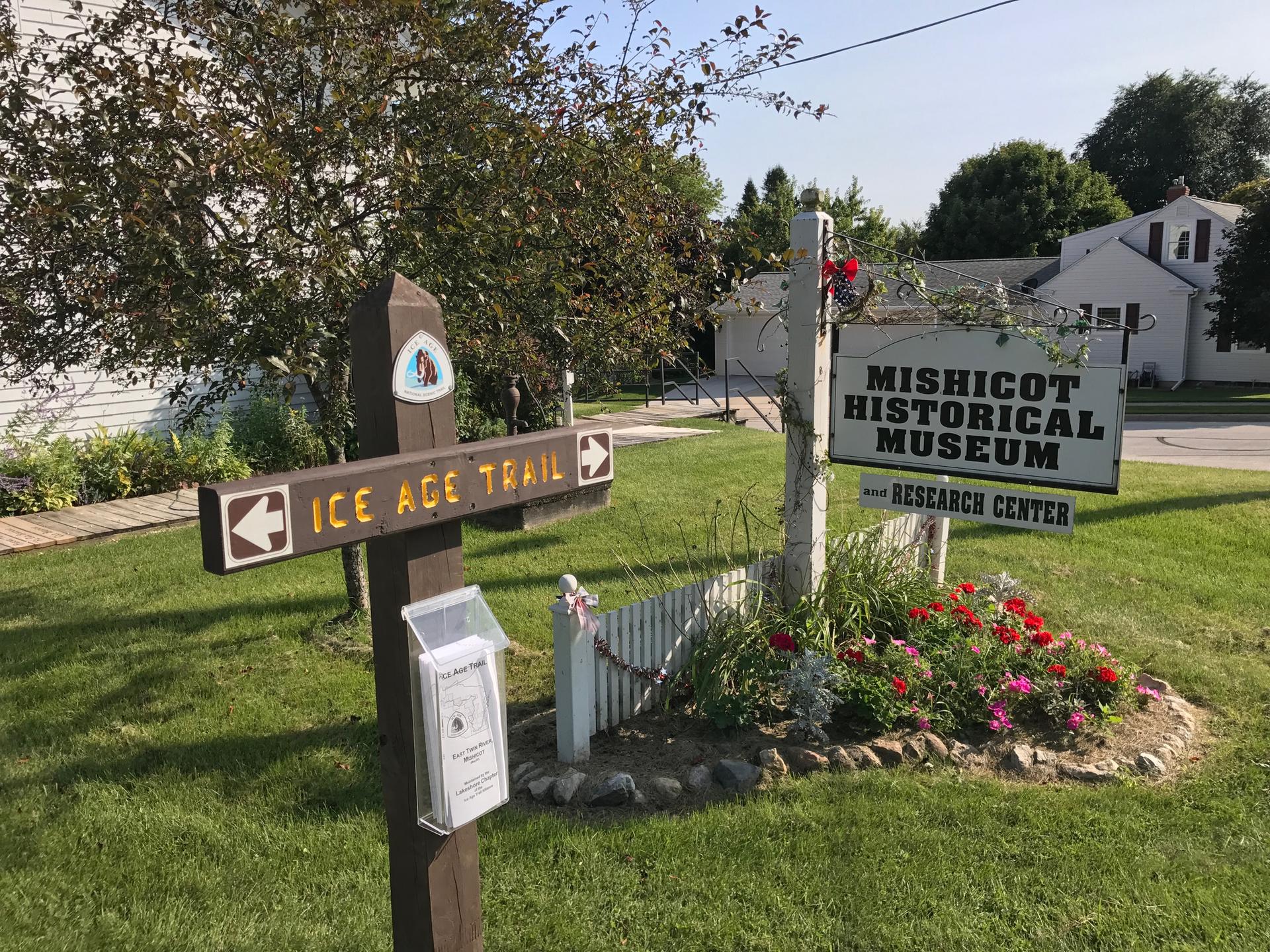 Two outdoor signs, one brown and one white, stand in a sunny, green landscape.