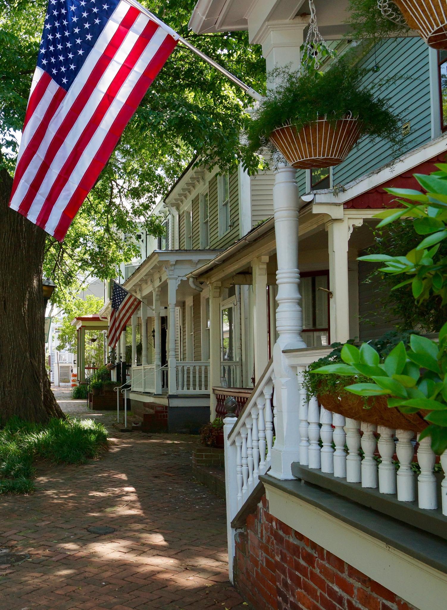 An American flag hangs from a porch of a historic house on a sunny day.