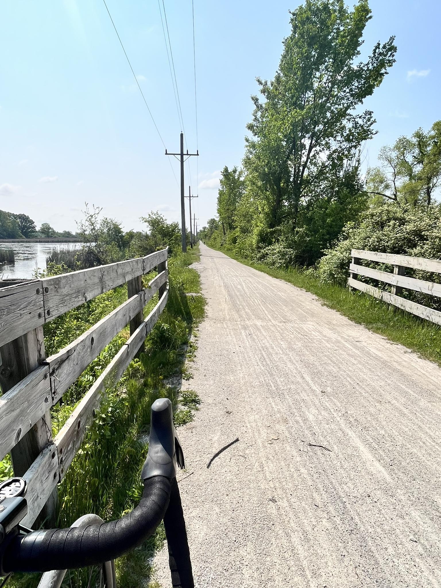 View from bicycle handlebars on a sunny dirt path next to a lake.